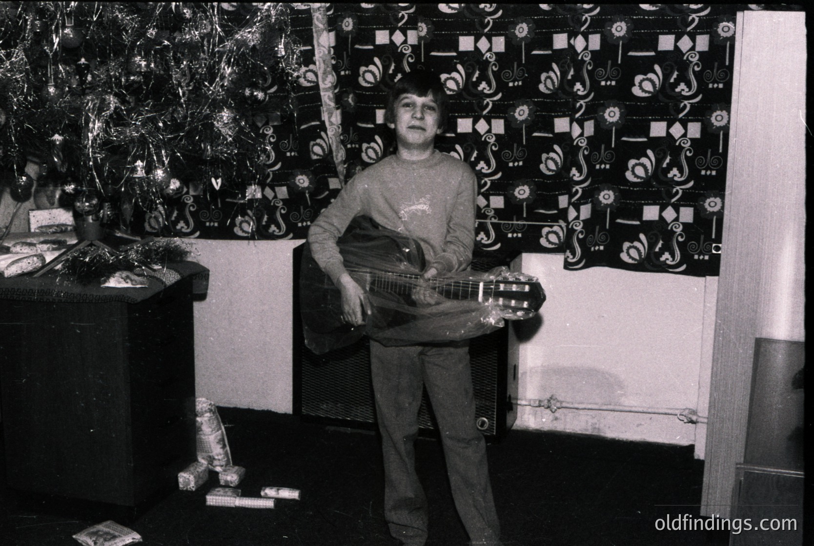 Young boy in 1970s-style sweater plays acoustic guitar indoors, framed by patterned wallpaper with geometric motifs. Decor includes a decorated Christmas tree and vintage fireplace. Candid, nostalgic home setting.