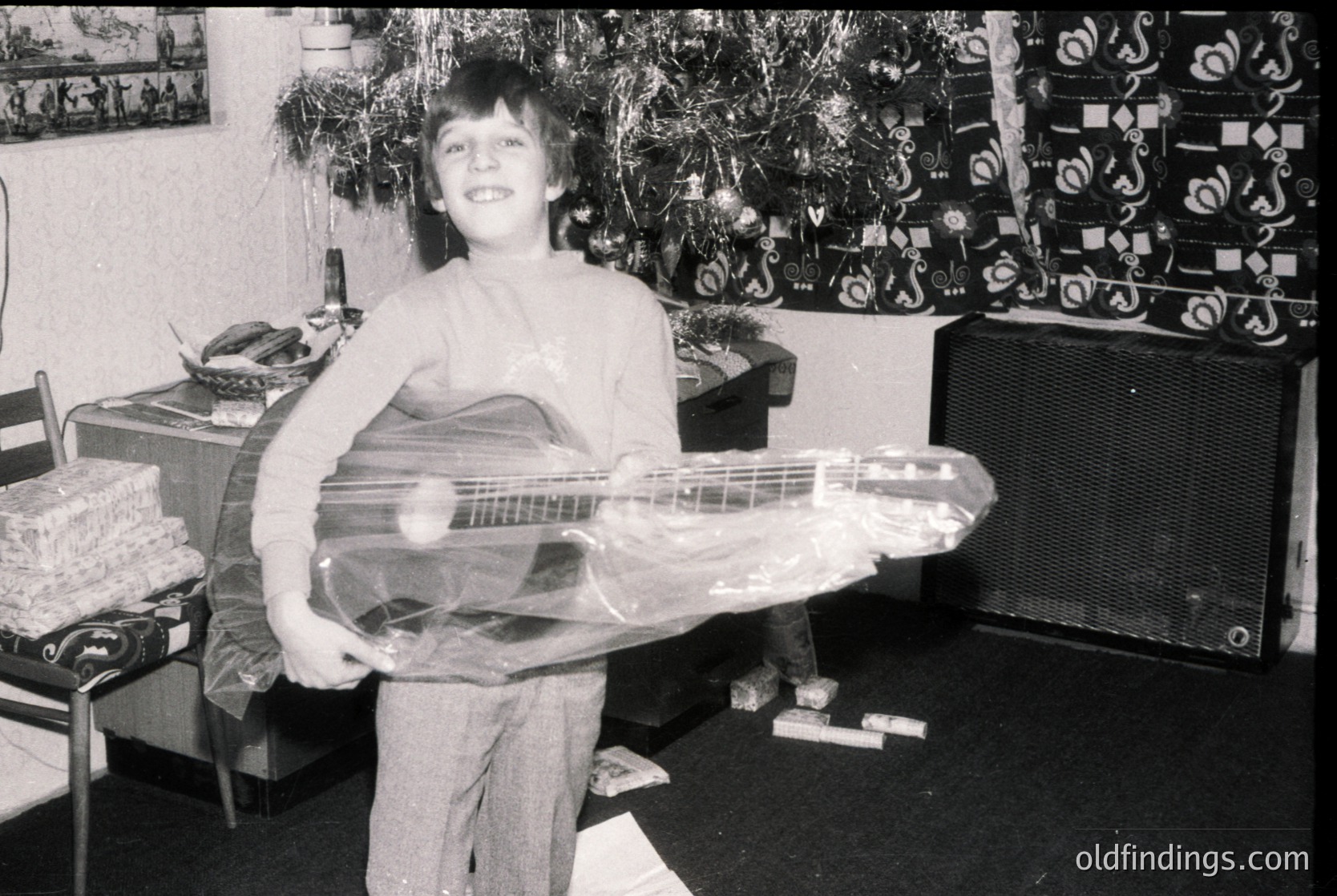 Young boy in 1970s-style sleeveless shirt and trousers poses indoors with a wrapped electric guitar, likely a gift. Surroundings include a decorated Christmas tree, vintage wallpaper, and a large speaker. Classic mid-century home setting.