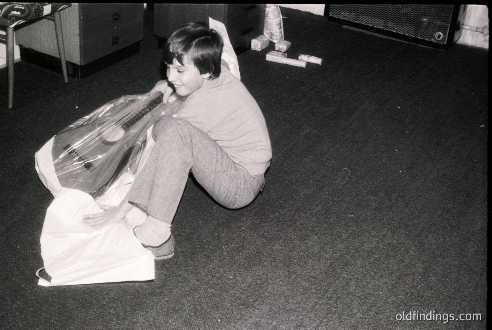 Child playing with a large plastic bag, possibly mimicking a guitar or musical instrument, in an indoor setting. Mid-20th century (1960s–1970s) domestic environment with vinyl record player in background. Casual, playful moment capturing youthful creativity.