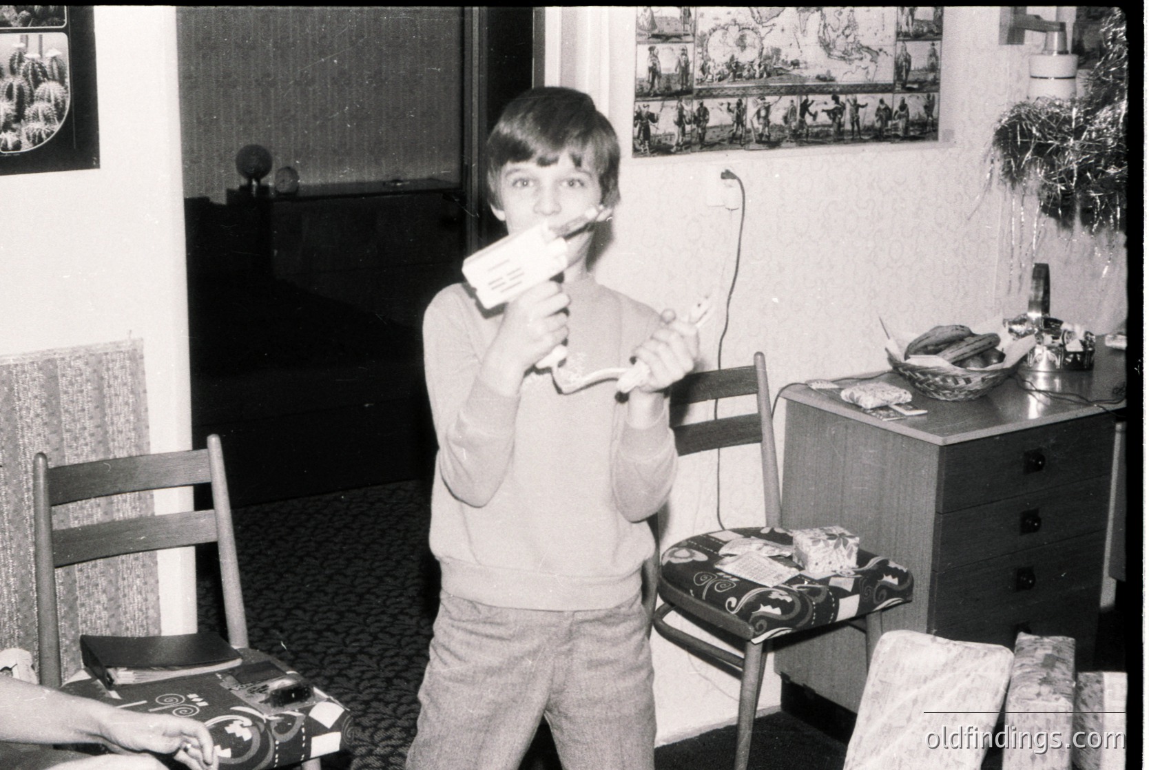 Black-and-white indoor shot of a young boy (mid-1970s) holding a toy gun and a toy sword in a mid-century living room. Wooden furniture, patterned carpet, and framed artwork on walls. Classic retro home decor with a vintage dresser and chair.