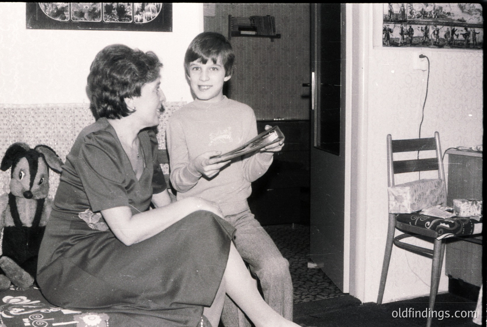 A woman and child in a mid-20th-century living room, likely 1960s–1970s. Woman in a short-sleeve blouse and skirt holds a book, engaging with the child seated beside her. Framed photos on walls depict outdoor scenes. Plain wooden chair and basic furnishings suggest modest domestic life.