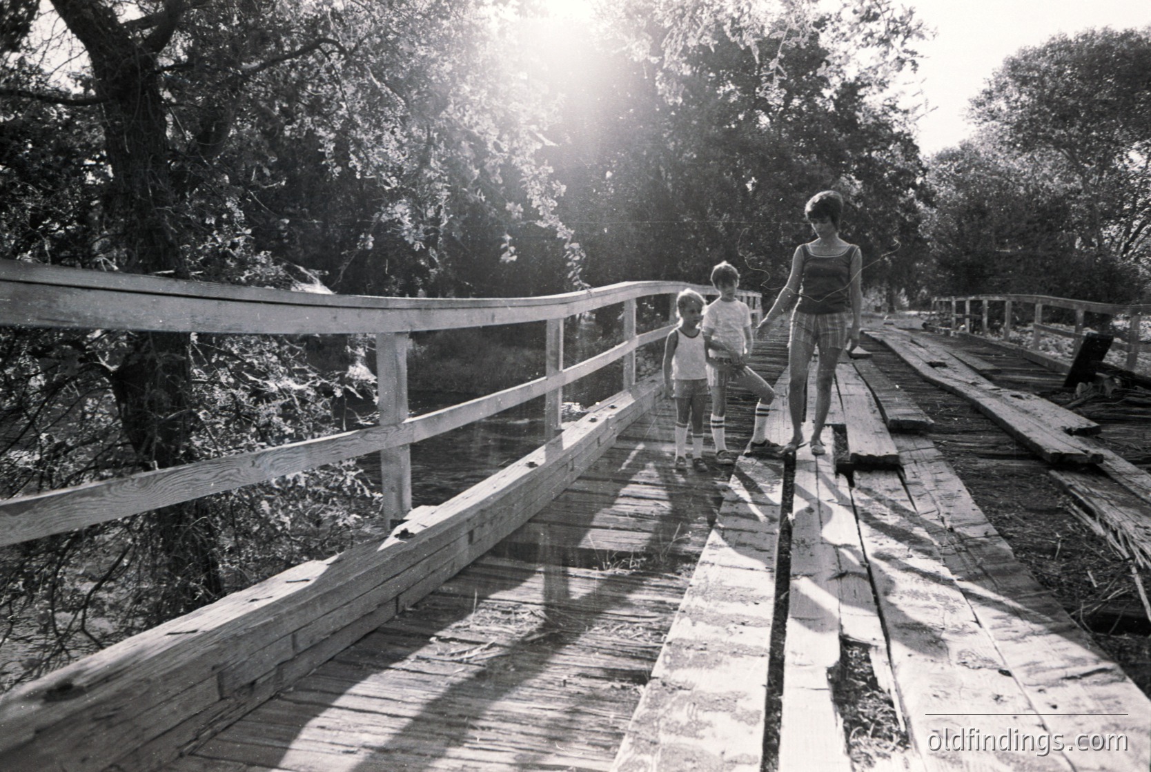 Three children cross a rustic wooden bridge in a sunlit forest, framed by dense foliage. The bridge’s weathered planks and handrails suggest a rural or park setting, likely mid-20th century. The warm sunlight and natural lighting evoke nostalgia.