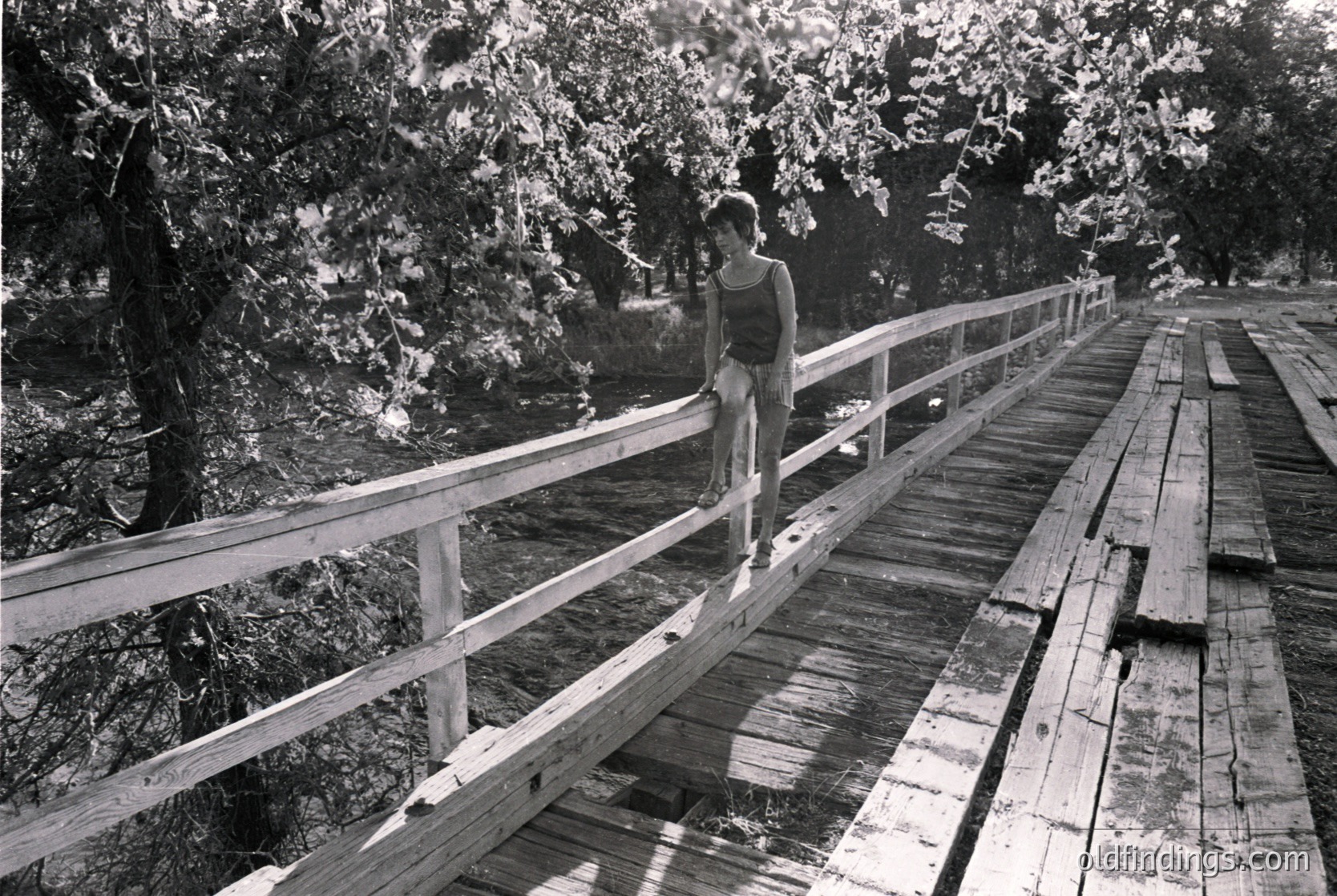 A lone individual stands on a weathered wooden boardwalk, leaning against a metal railing. The scene captures mid-20th century outdoor leisure, likely 1950s–1960s. Lush greenery and dense foliage frame the path, suggesting a park or nature reserve. The person’s casual attire and relaxed posture evoke a sense of exploration and freedom. [Mid-20th-century boardwalk with lone figure against metal railing in lush greenery ]