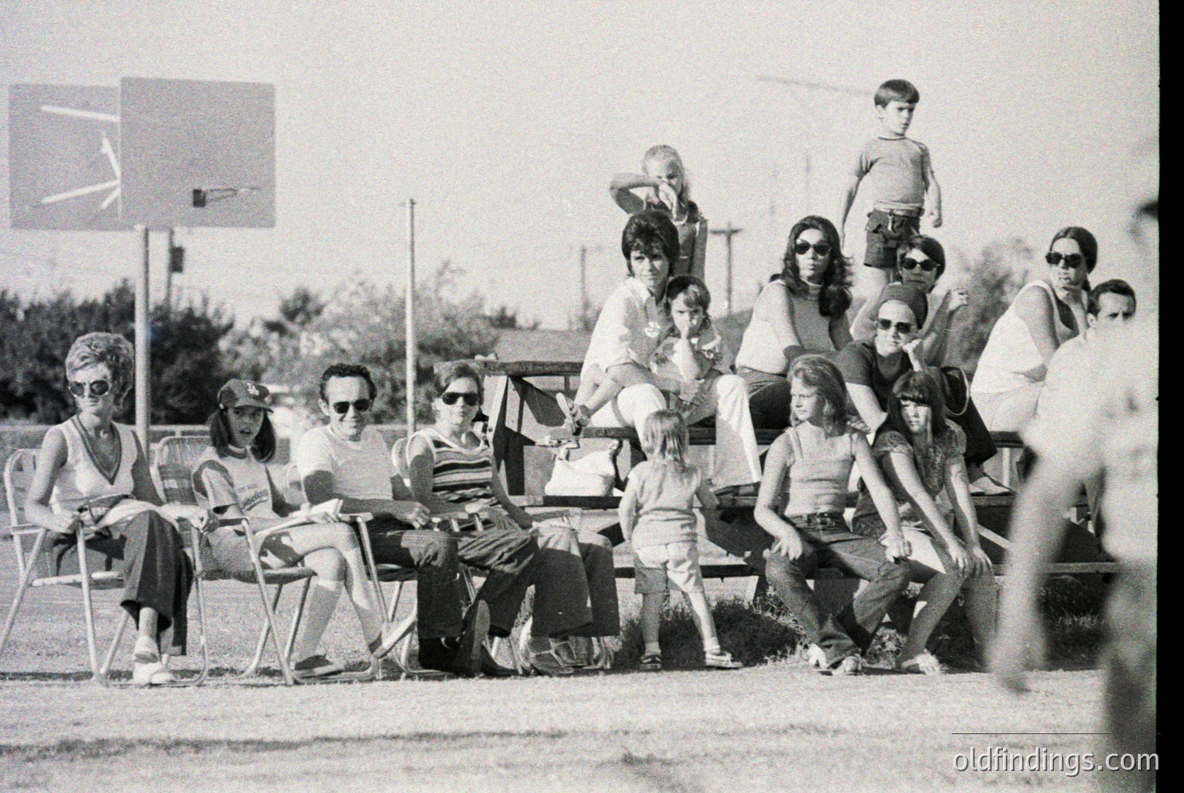 Black-and-white snapshot of a mid-20th-century outdoor gathering, likely 1960s–1970s. Group of ~15 people—adults and children—sitting on bleachers and benches near a basketball hoop. Casual attire: striped shirts, sunglasses, and bell-bottoms. One child stands atop bleachers. Background shows grassy field and residential houses.