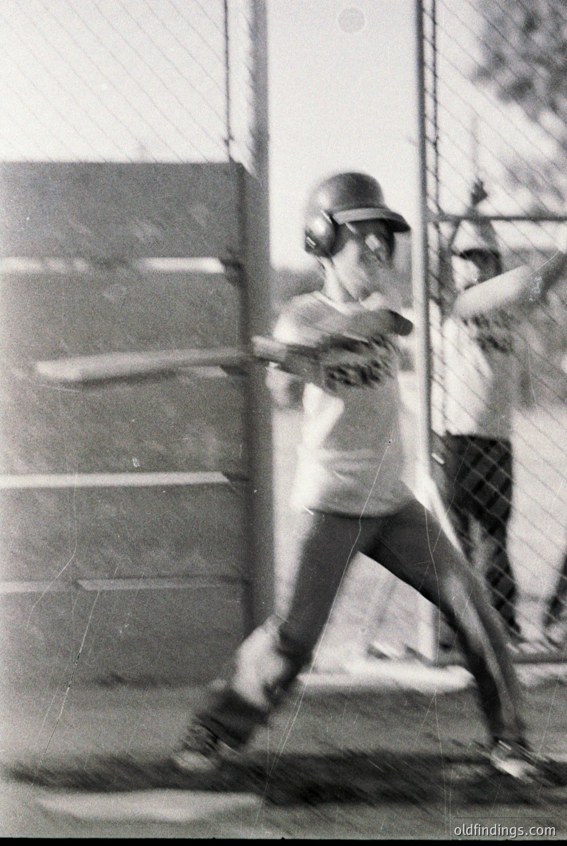 Black-and-white photo of a baseball player mid-swing in a batting cage, wearing a helmet and protective gear. Cage mesh and concrete steps visible in background. Likely 1960s–1980s American youth sports setting.