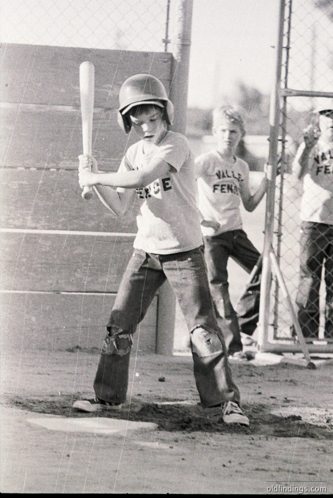 Young baseball player in 1970s-era protective gear—helmet, knee pads, and belt—mid-swing at a school or youth league game. "Valley Feeder" jersey suggests a team affiliation. Graffiti on concrete wall ("SEX") and dirt infield highlight informal, community setting.