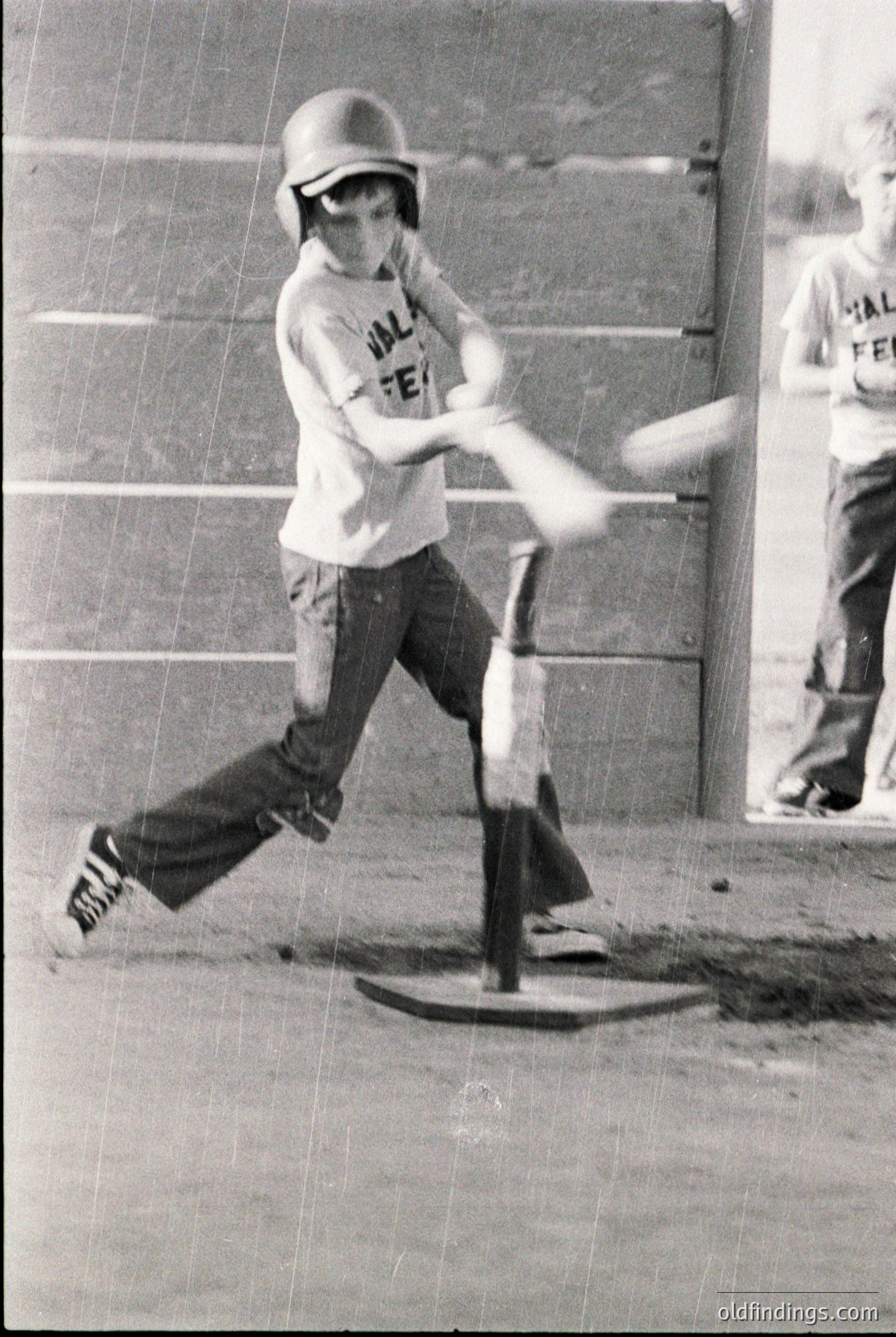 Young baseball player mid-swing in vintage helmet and "Valley Feeder" jersey, 1960s-70s. Indoor concrete court with T-shaped batting tee. Motion blur captures dynamic follow-through.