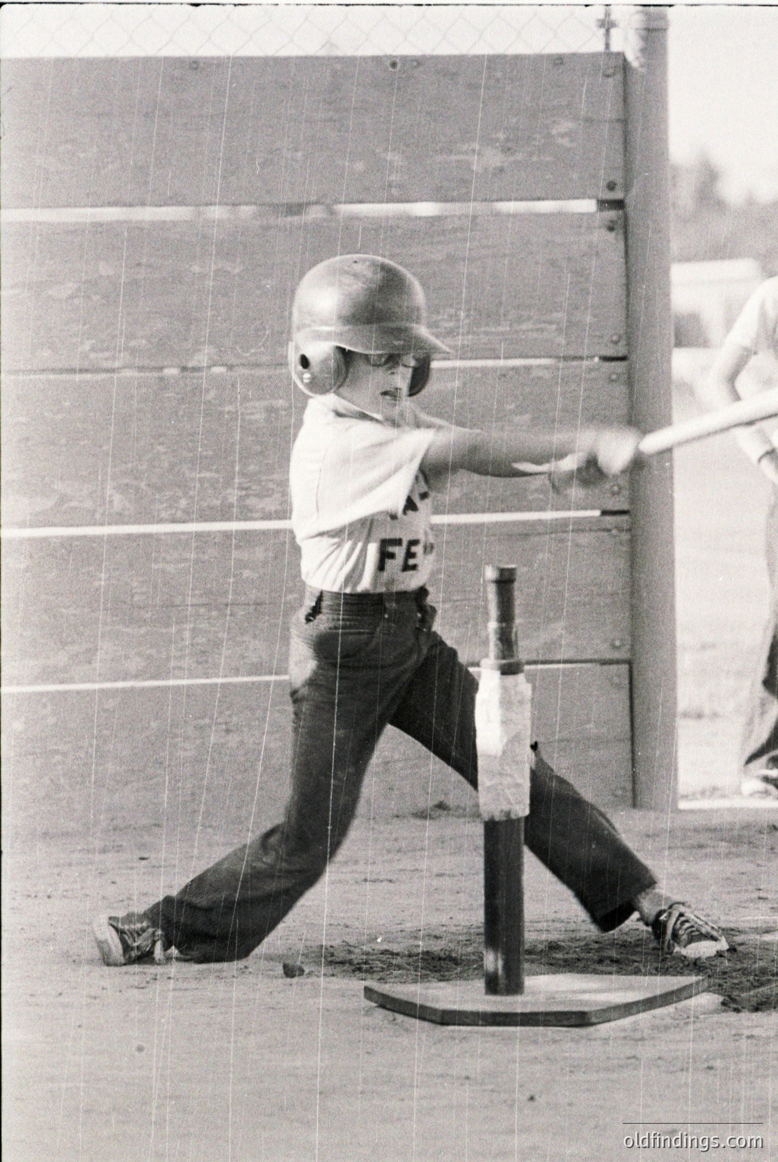 Young baseball player mid-swing in vintage batting helmet and "FE" jersey, 1960s-70s style. Indoor batting cage with wooden fence and tee setup. Classic youth sports action shot.