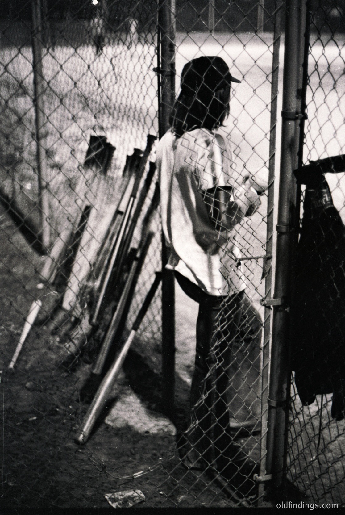 Baseball player in vintage uniform stands at dugout entrance, gripping bat. Chain-link fence and dugout bench visible. Likely 1950s–1970s American minor league setting.