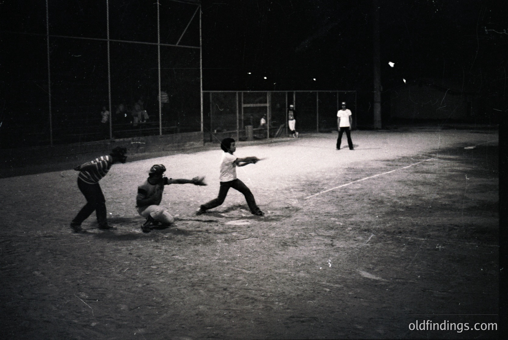 Mid-20th century baseball game under stadium lights, featuring three players in action: batter, catcher, and infielder. Concrete bleachers and glass-paneled walls in background suggest urban or school setting. Monochrome film captures dynamic motion and vintage sports culture.