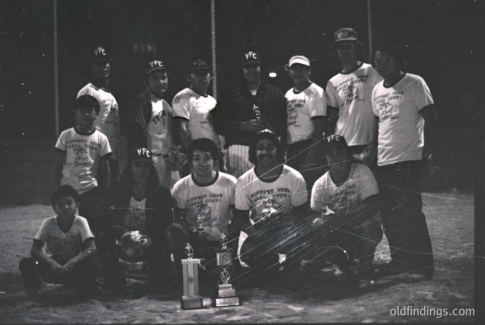 YFC baseball team poses with championship trophy, 1970s. Indoor stadium setting with visible net and lighting. Uniforms feature "YFC" logos and "Support Your Church" text.