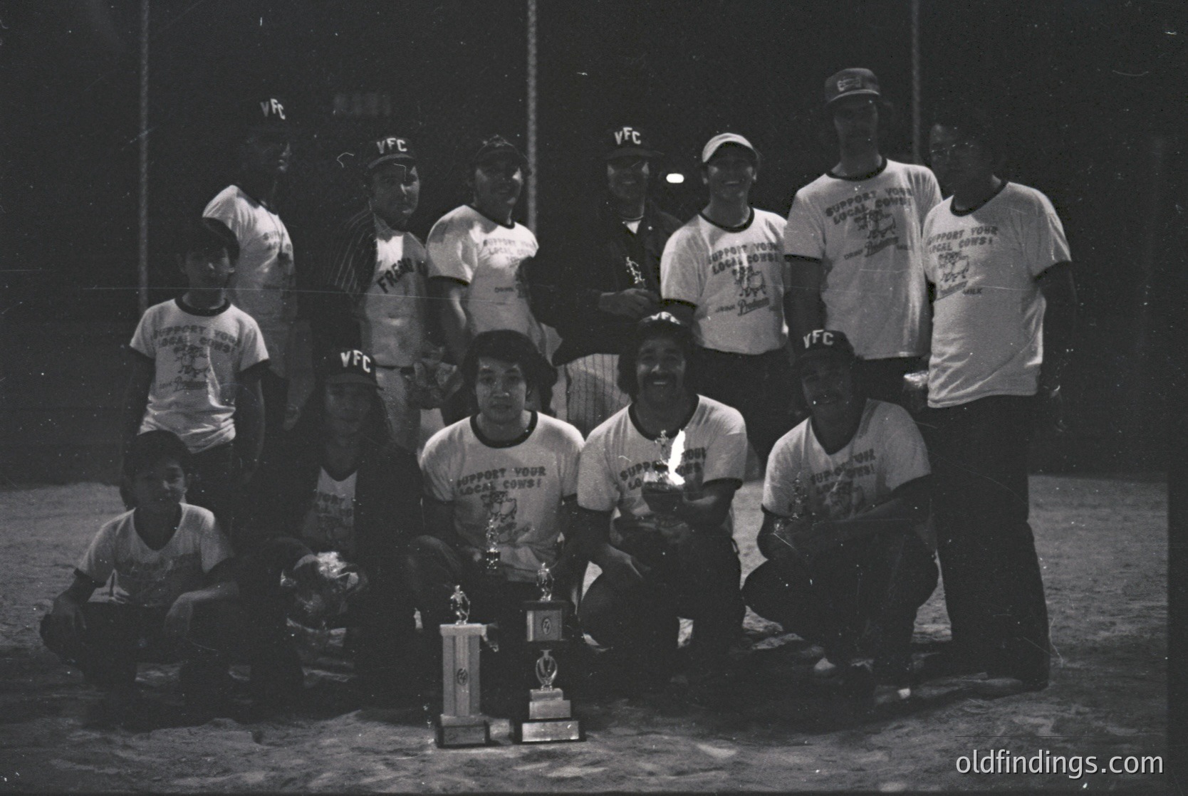 Vintage black-and-white photo of a youth football team posing with a trophy on a dirt field. Uniforms feature "YFC" logos and "Support Your Community" text, suggesting 1970s–1980s era. Backdrop includes a chalkboard with "YFC" markings. Ideal for sports history or community sports research.
