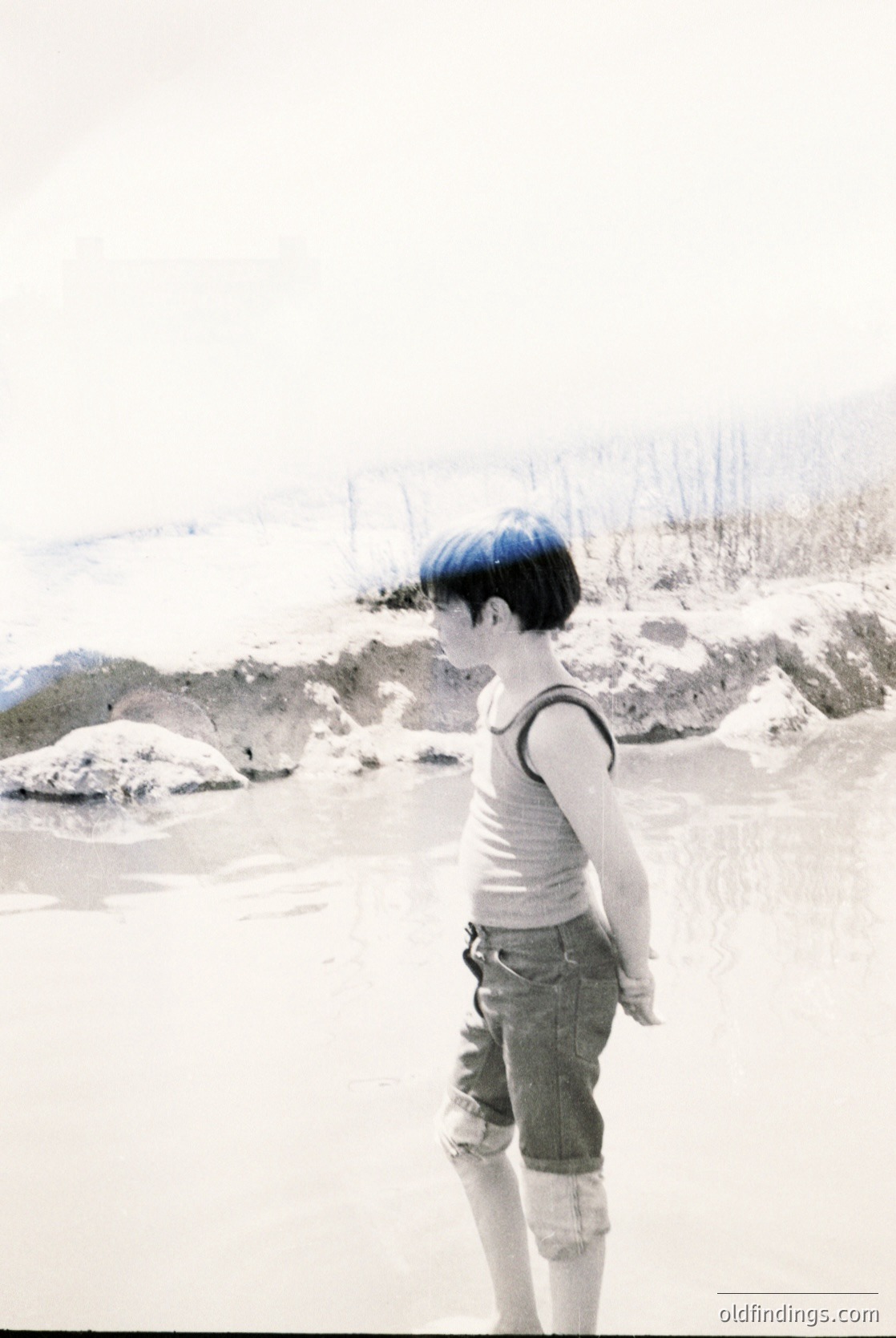 Child with blue-dyed hair stands on a snowy, rocky shore, facing water. Mid-20th century street fashion: sleeveless white top, rolled denim shorts, and knee-high socks. *(Note: If this is a vintage photo, consider adding or for broader searchability.)*