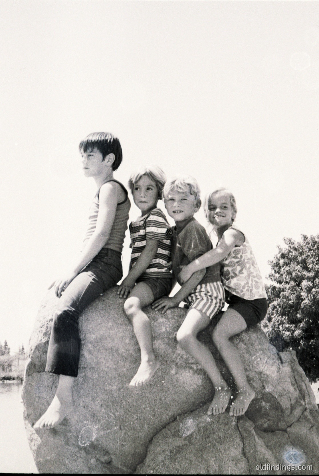 Four children pose playfully on a large rock by water, mid-20th century. Boys in striped swim trunks and tank tops, girls in sleeveless dresses, all barefoot. Classic summer attire reflects 1950s–1960s beach culture.