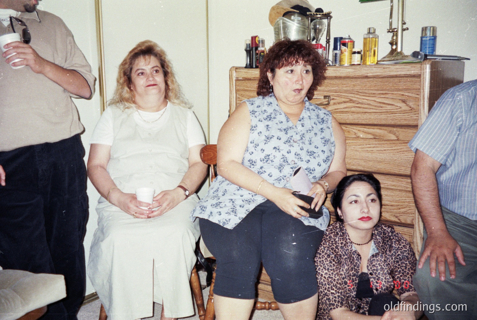 Vintage indoor gathering featuring four adults in a modestly furnished room, likely late 20th century. Central woman in floral top and black shorts holds a white object, surrounded by others in casual attire. Wooden dresser with metalware and bottles in background. Warm, lived-in atmosphere.