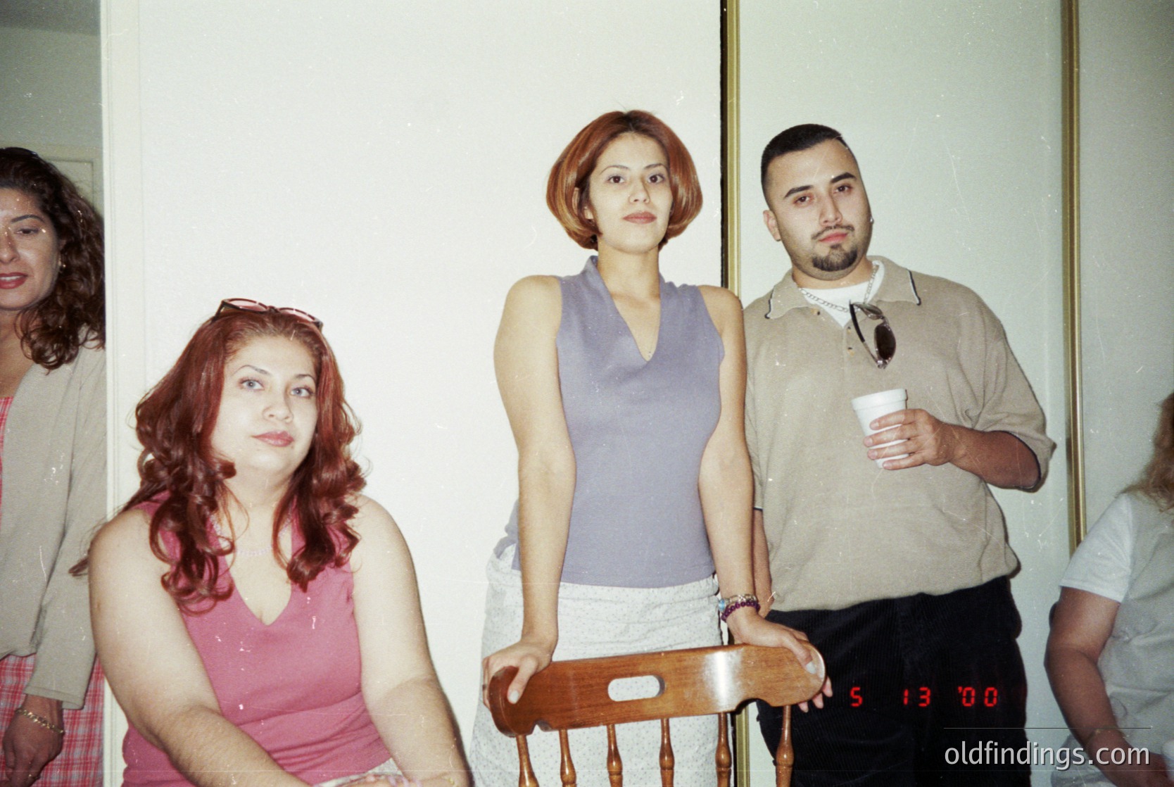 Vintage group portrait in indoor setting, likely 1990s–2000s. Three central figures pose: woman in sleeveless purple top, man in beige shirt holding coffee cup, and woman in pink tank top. Wooden chair with visible "S 13'00" label in foreground. Soft lighting, muted colors, and slight grain suggest analog photography. Potential cultural context: social gathering or informal event.