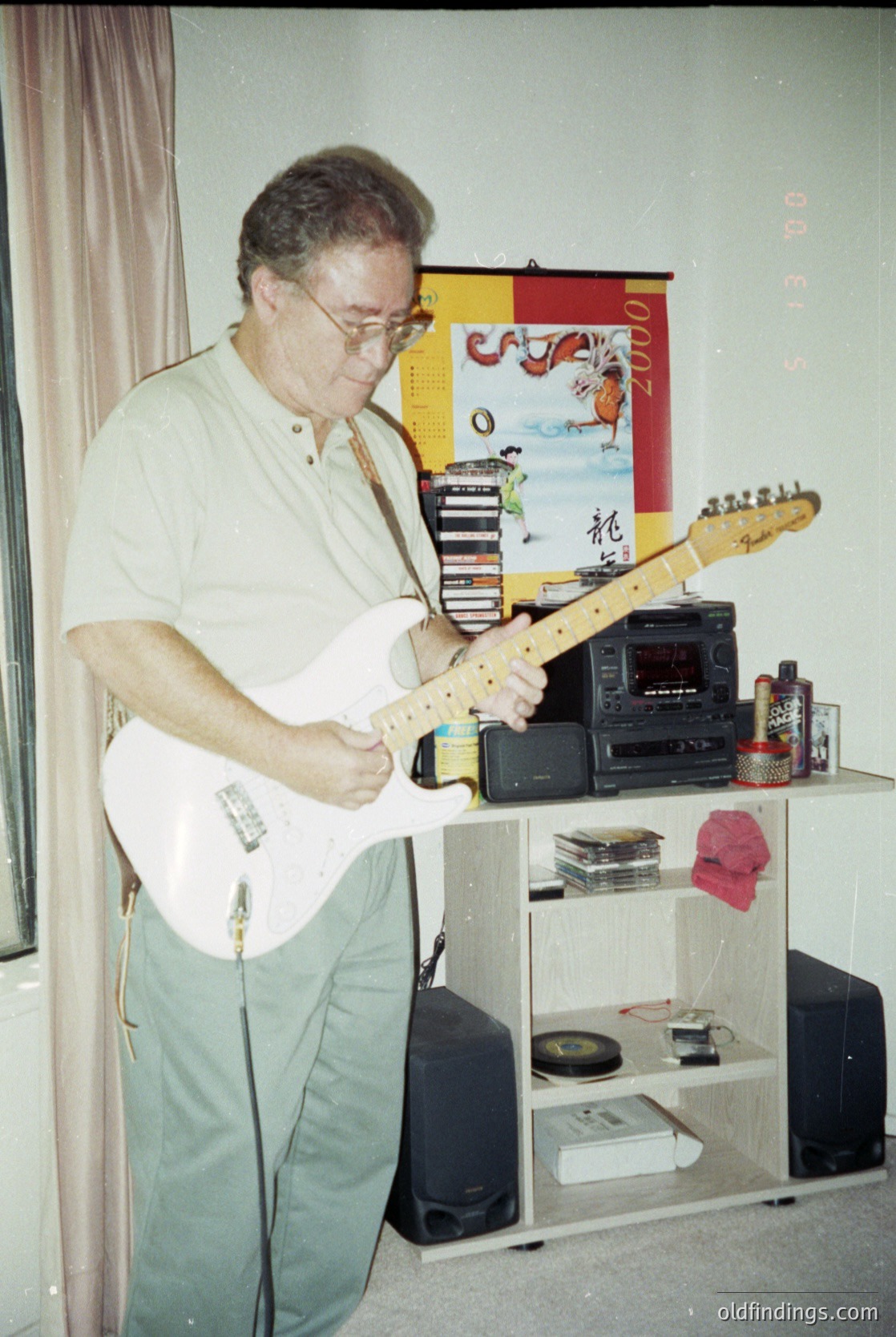 Mid-century indoor setting featuring a man playing a sunburst-toned electric guitar, likely a Fender-style model. Surrounding him: a cassette tape player, vinyl records, and a framed decorative banner with Asian-inspired motifs. Lighting suggests indoor evening or studio ambiance. Potential 1970s–1980s timeframe.