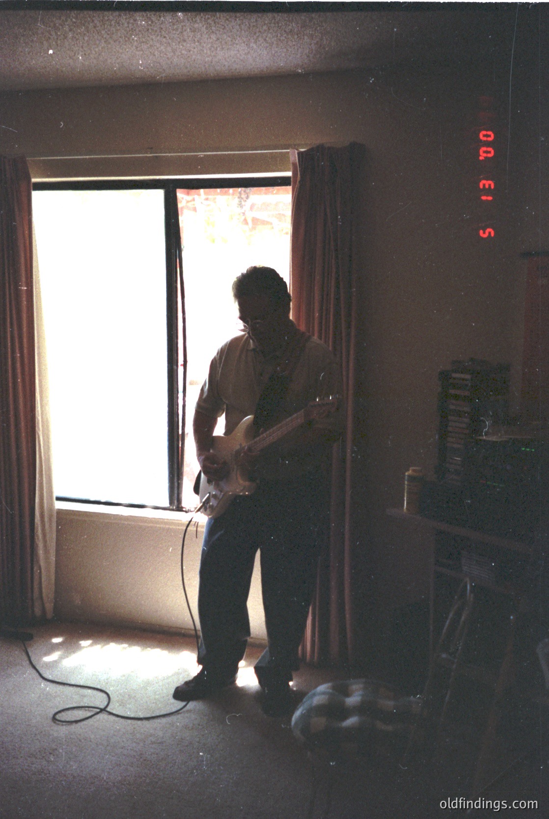 Mid-century guitar studio with analog clock (5:13 PM) showing time. Man in casual 1960s–70s attire plays electric guitar near large window. Stacked vinyl records on shelf, vintage radio receiver on desk. Warm lighting highlights retro interior.