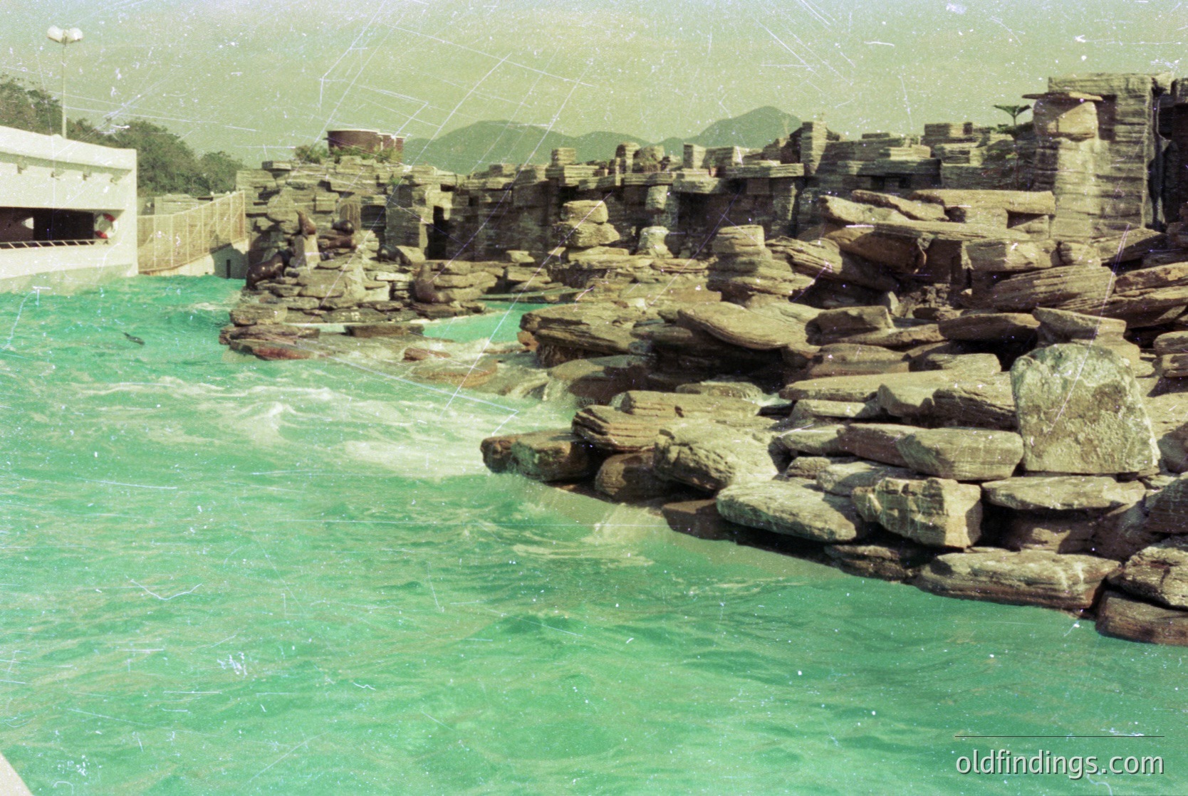 Rustic stone breakwater with stacked boulders protecting a coastal structure, likely a 1970s seaside resort. Green-tinted water contrasts with aged concrete and weathered rocks.