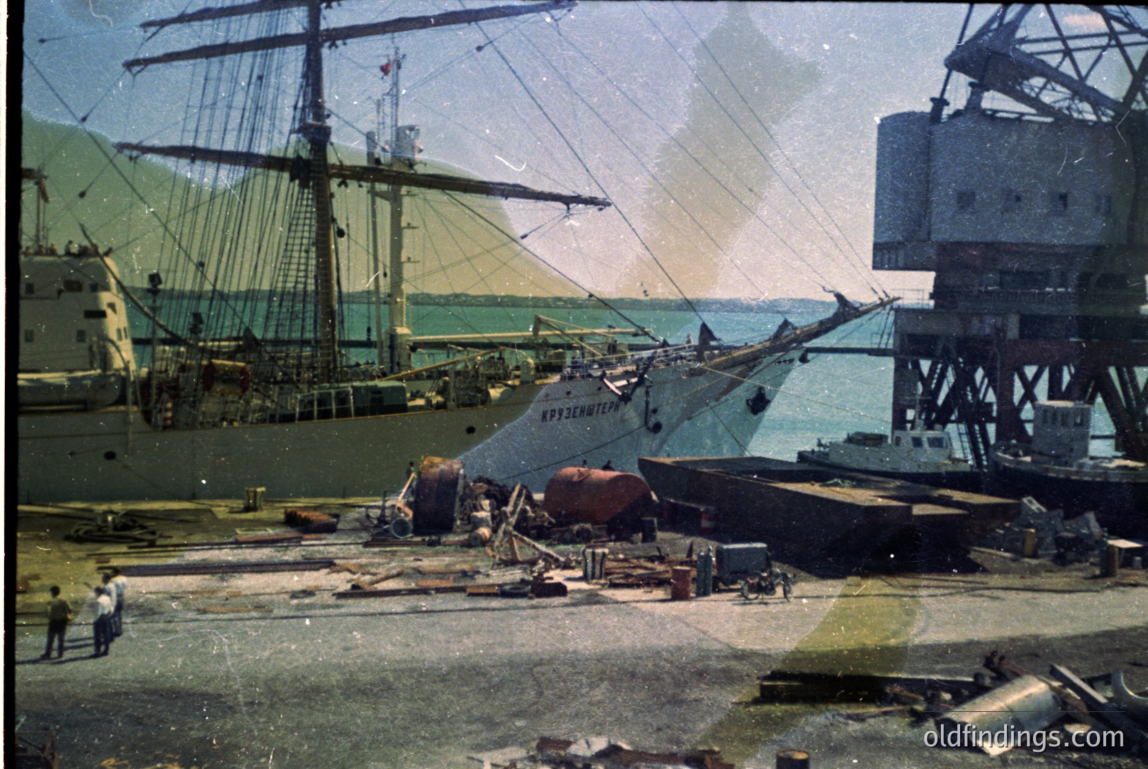Vintage sepia-toned shipyard scene with a multi-masted sailing vessel docked alongside industrial cranes and scaffolding. Foreground shows debris and workers in early 20th-century attire. Likely a European port, possibly Baltic or North Sea region.