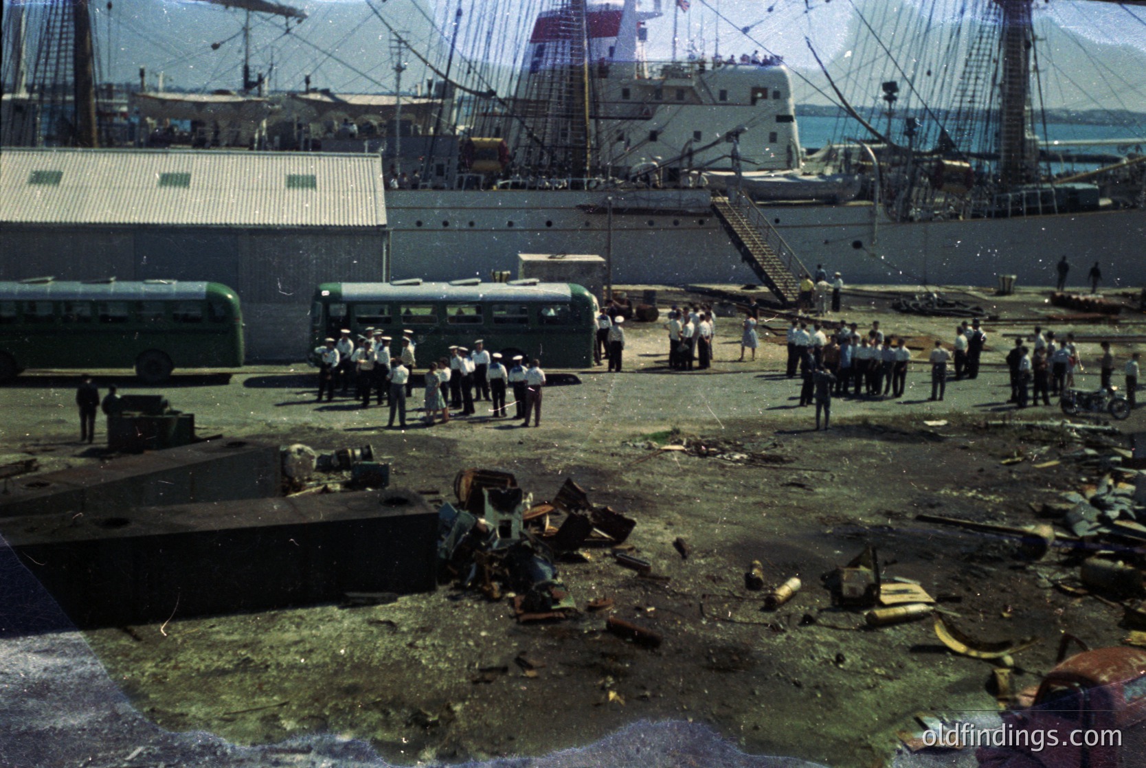 Vintage port scene with industrial docks, mid-20th century. Workers in uniform stand near debris and cargo, with a large ship and smaller bus in background. Likely Eastern Bloc port infrastructure, 1950s-1960s.
