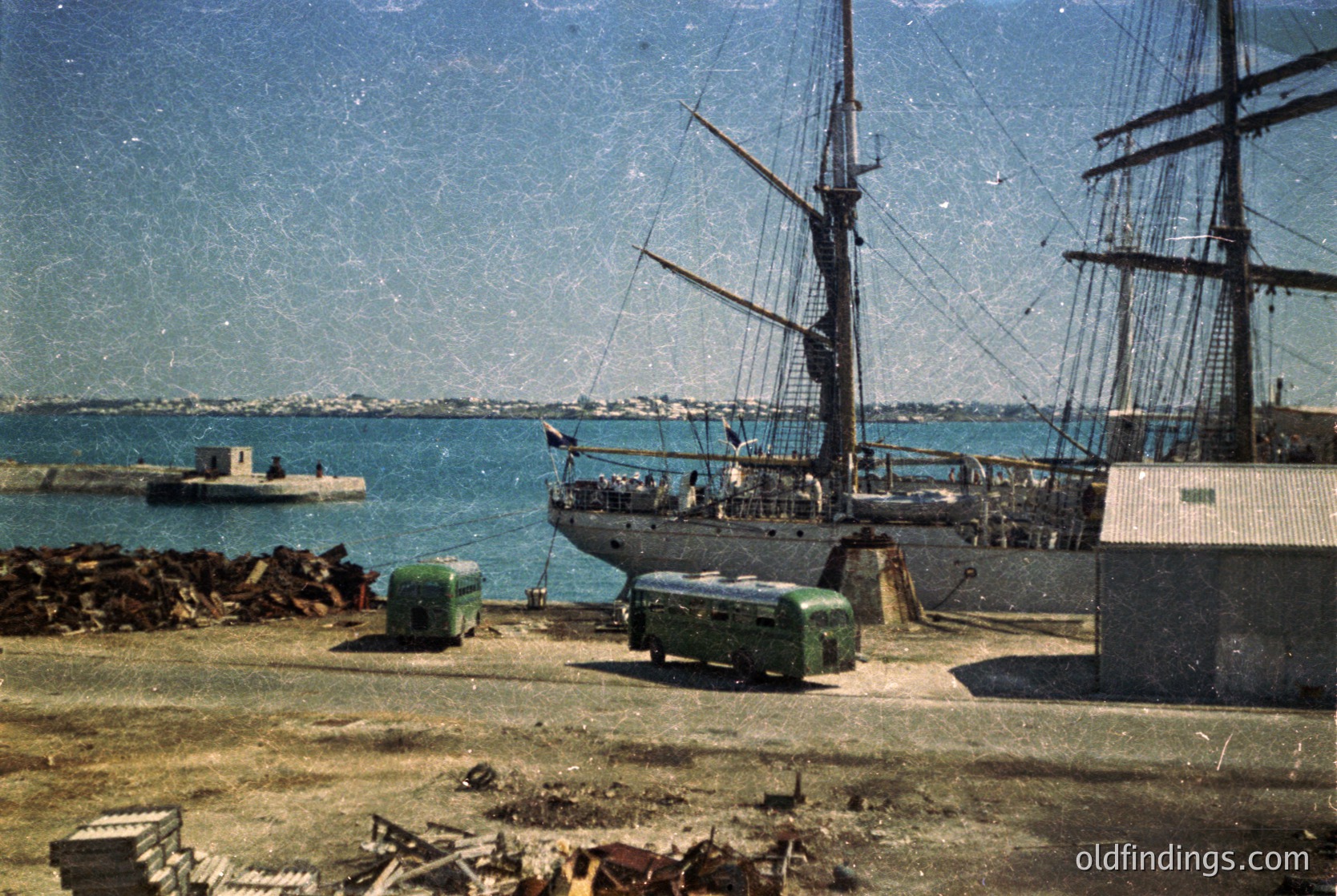Vintage seaside dock with a wooden sailing ship moored alongside. Green industrial containers and stacked lumber in foreground. Coastal cityscape in background under clear skies. Likely mid-20th century maritime port.