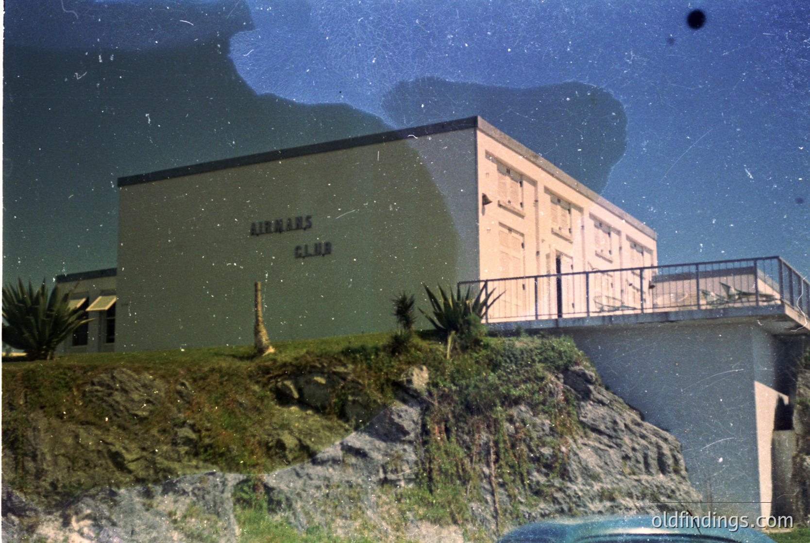 Mid-century seaside building labeled "Airman’s Club" perched on rocky terrain with concrete steps and railings. Green exterior with white accents, palm plants, and a balcony overlooking the sea. Likely a 1950s–1960s military or resort facility.