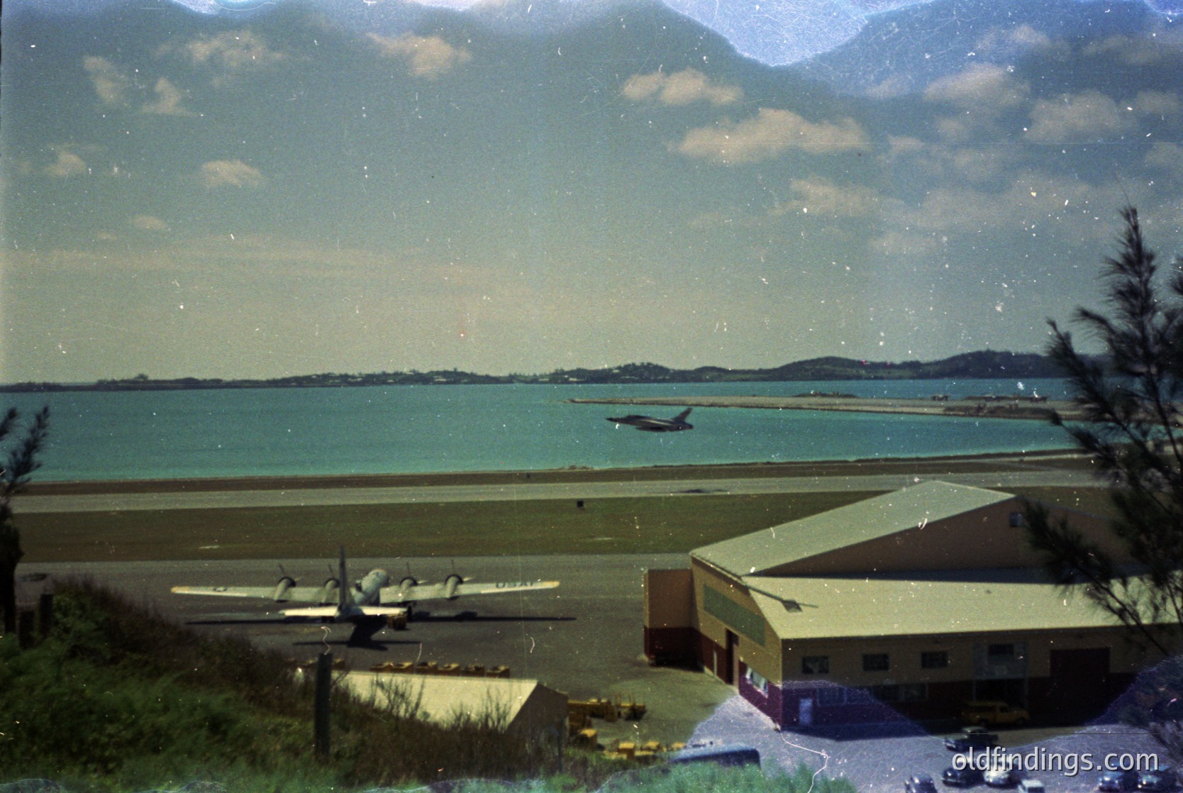 Vintage aerial view of a coastal airfield with twin-engine propeller aircraft on a grass runway. Mid-20th century hangar and small terminal building in foreground. Distant island and calm sea under overcast skies.