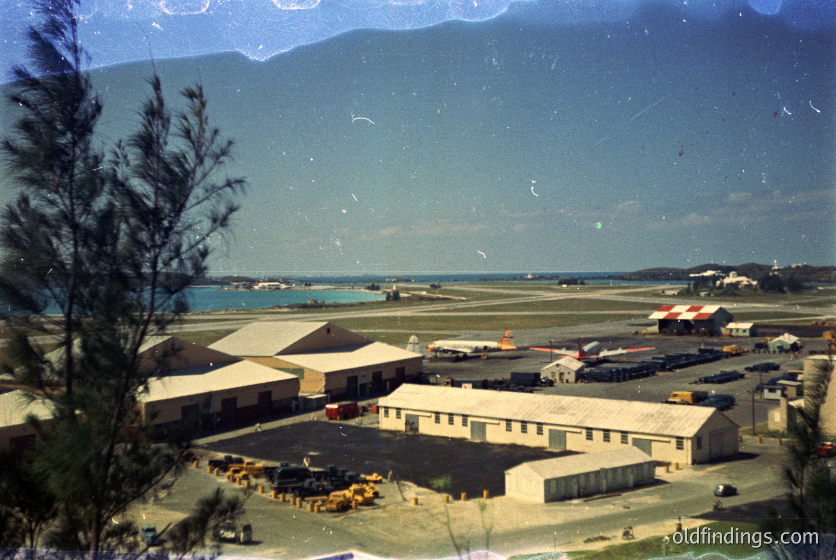Vintage aerial view of a coastal airfield with mid-century concrete buildings, parked aircraft, and vehicles. Mountain range and ocean backdrop suggests a tropical or subtropical location. Likely 1950s–1970s era.