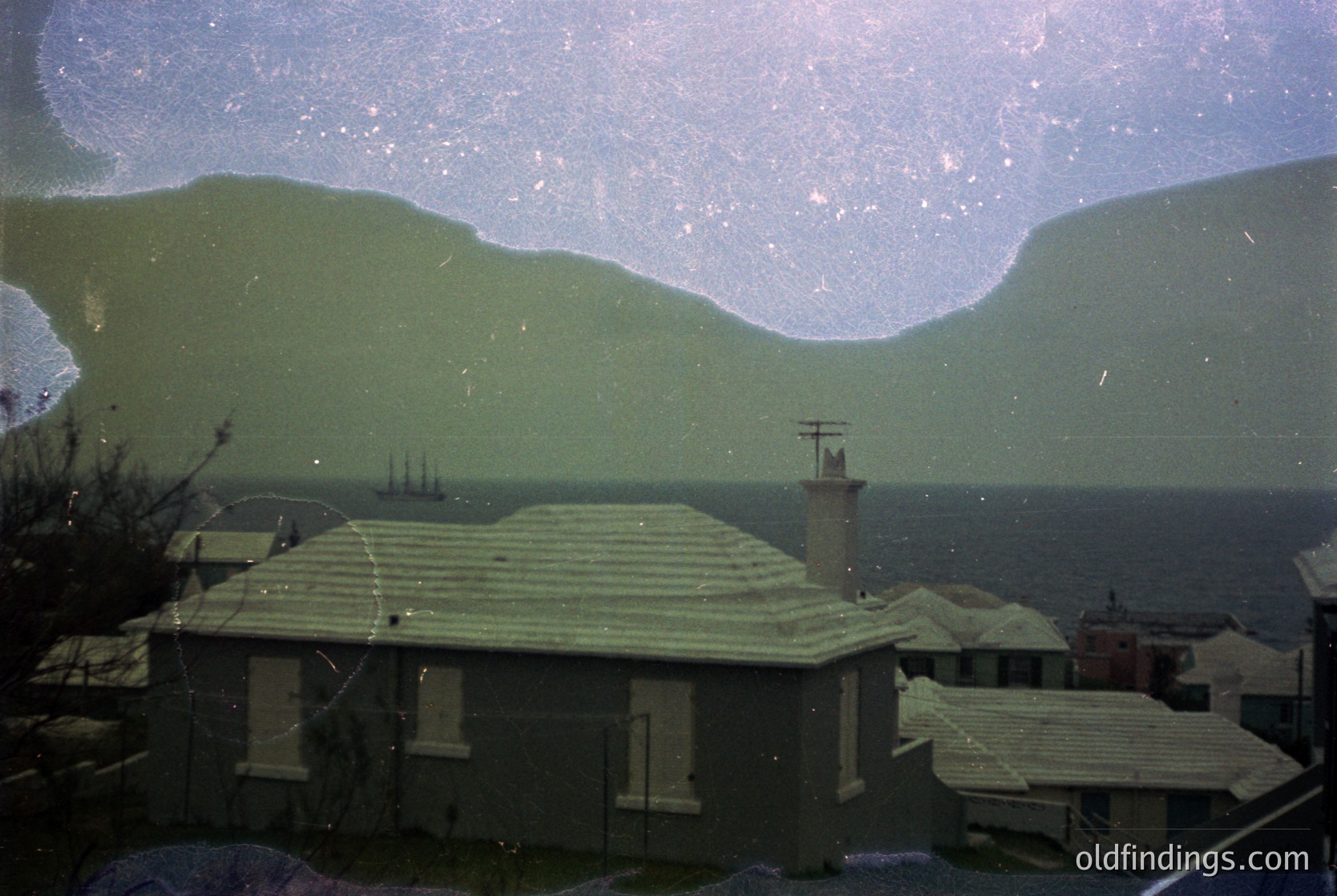Vintage coastal scene through aged glass—distorted but revealing: low-slung beach houses with flat roofs, a chimney, and utility wires. Distant ship silhouetted against misty sea and rolling hills. Likely Mediterranean or Adriatic region, 1950s–1970s.