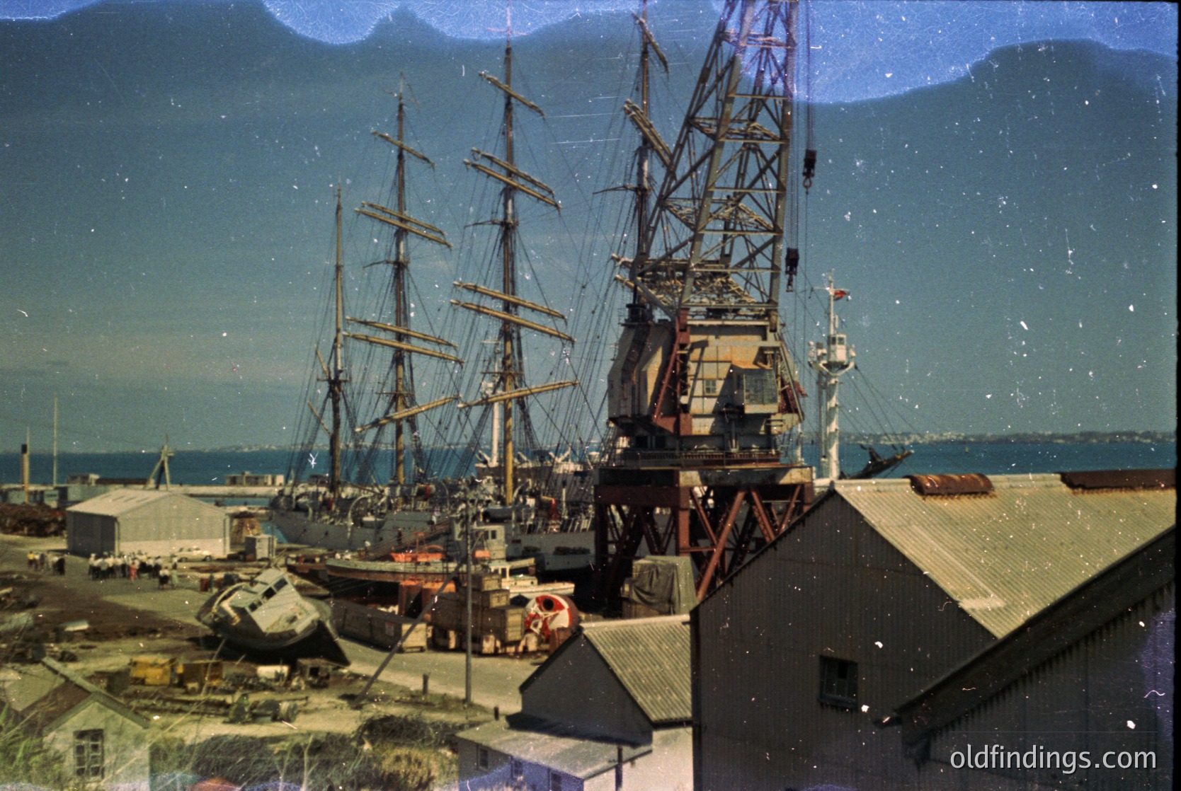 Vintage aerial view of a dockyard featuring a large **four-masted schooner** and industrial cranes, likely mid-20th century. Industrial buildings and a coastal landscape with mountains in the background suggest a maritime port. The grainy, sepia-toned filter enhances historical authenticity.