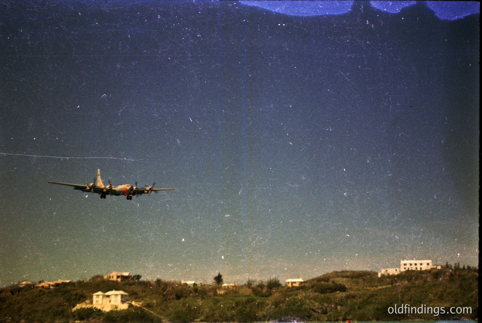 A vintage propeller aircraft ascends at night, illuminated by distant city lights against a star-studded sky. Likely a 1950s–1960s era transport plane, silhouetted against a horizon of low-rise buildings and greenery.