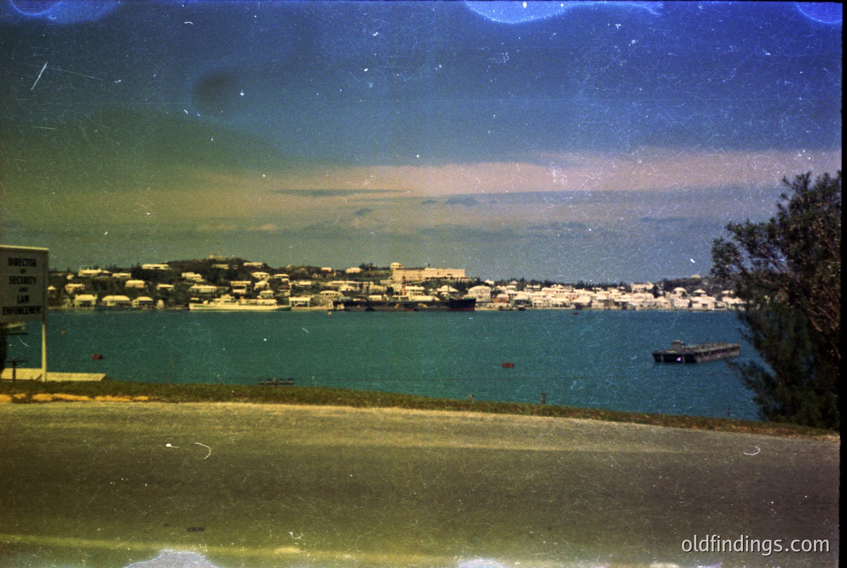 Vintage seaside town with low-rise buildings along a calm coastal road, captured through a slightly yellowed filter. Dense foliage frames the left edge, while a lone boat floats in the turquoise water. Likely mid-20th century, possibly Mediterranean or Adriatic region.