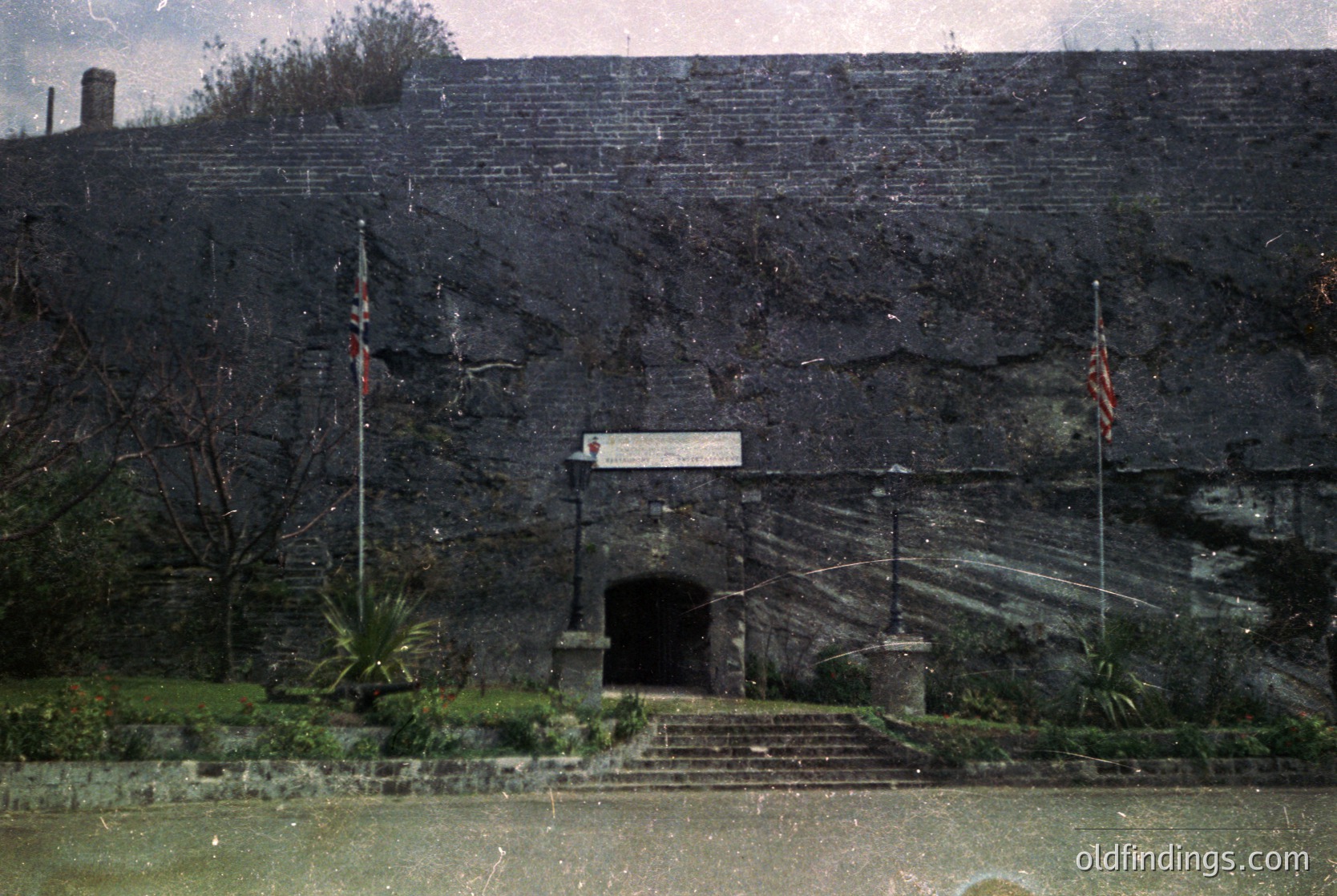 Historic stone entrance tunnel with arched doorway, flanked by two flagpoles. Surrounding vegetation includes sparse trees and shrubs. Likely a Cold War-era bunker or military structure.
