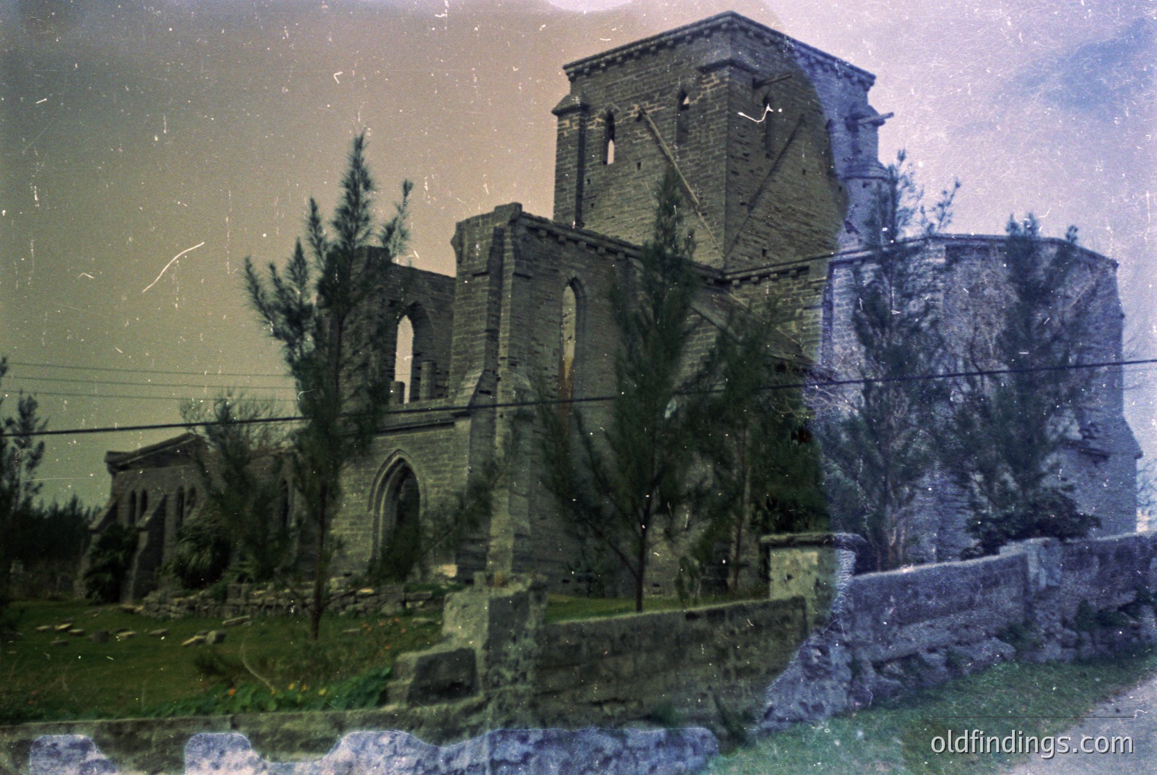 Vintage sepia-toned photo of a ruined Gothic-style stone church with arched windows and a bell tower, partially overgrown with greenery. Stone walls and a low boundary wall with rounded coping stones surround the site. Likely Eastern European, mid-20th century.