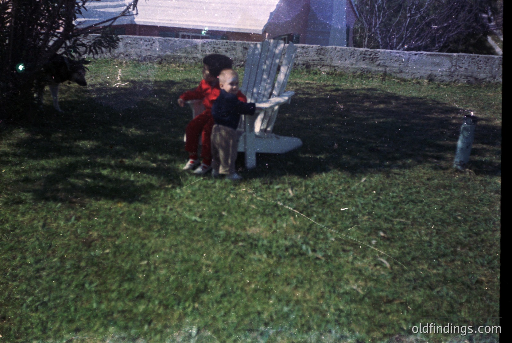 Two children in vintage clothing (1960s-70s) play on wet grass beside a metal bench. One wears a red coat, the other a striped sweater. A stone wall and house with a pitched roof frame the scene.