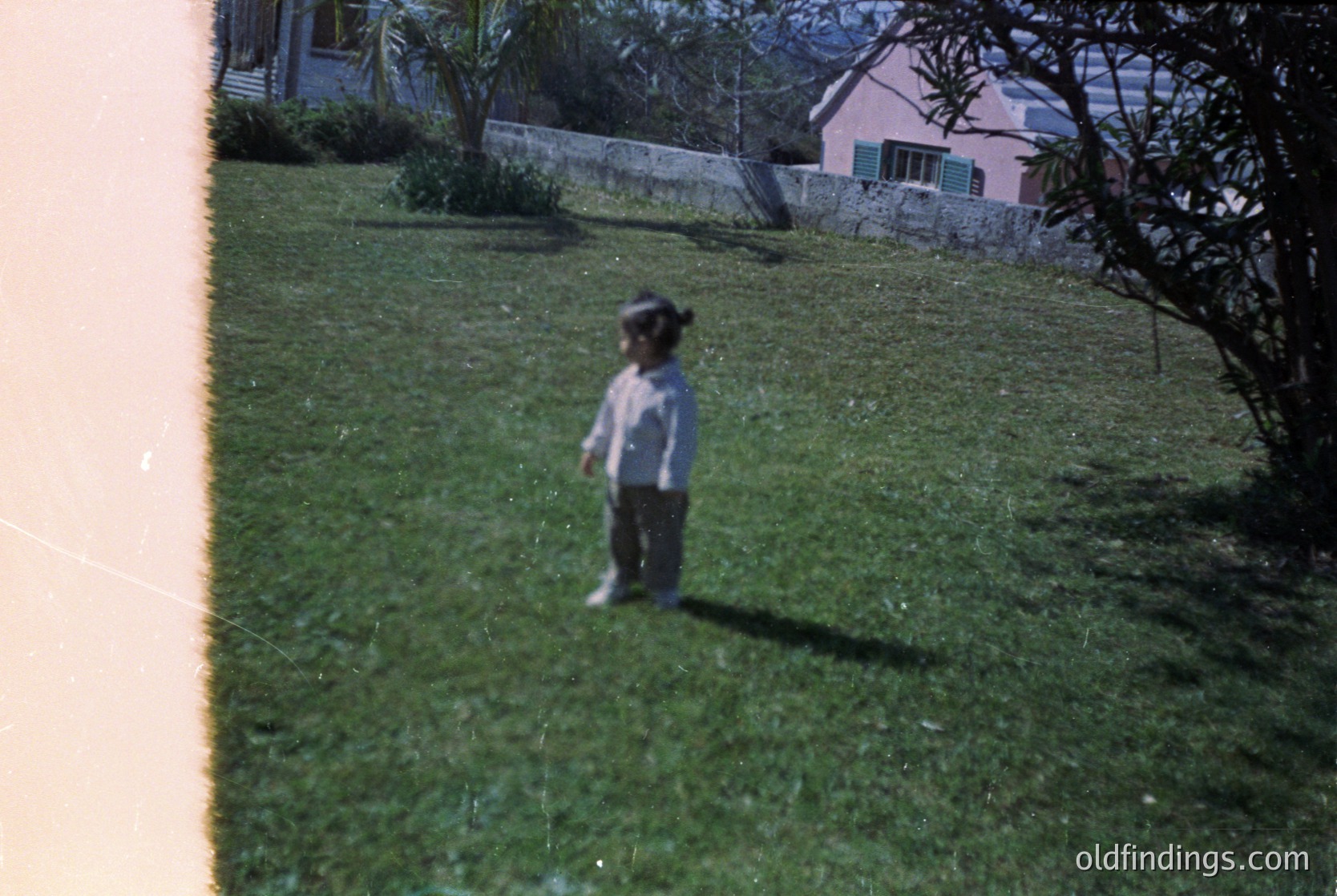 A child in mid-20th century attire stands on a well-manicured lawn, framed by a window edge. Lighting suggests vintage color processing (). Residential setting with pastel house and mature trees ( ) --- **Note:** Character count: 140. Tags reflect time period, setting, and archival relevance.