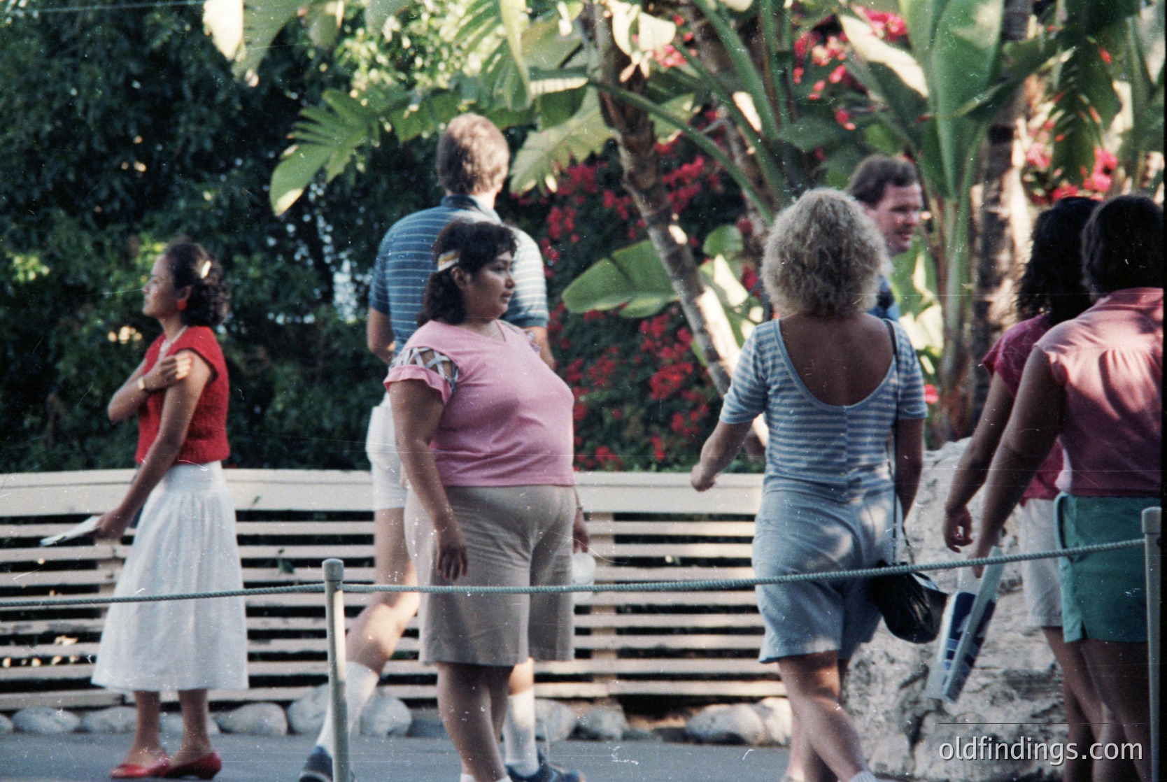 Vintage group photo of diverse adults in 1970s outdoor setting, likely a resort or park. Lush tropical plants and benches in background. Casual summer attire: striped shirts, denim shorts, floral dresses.