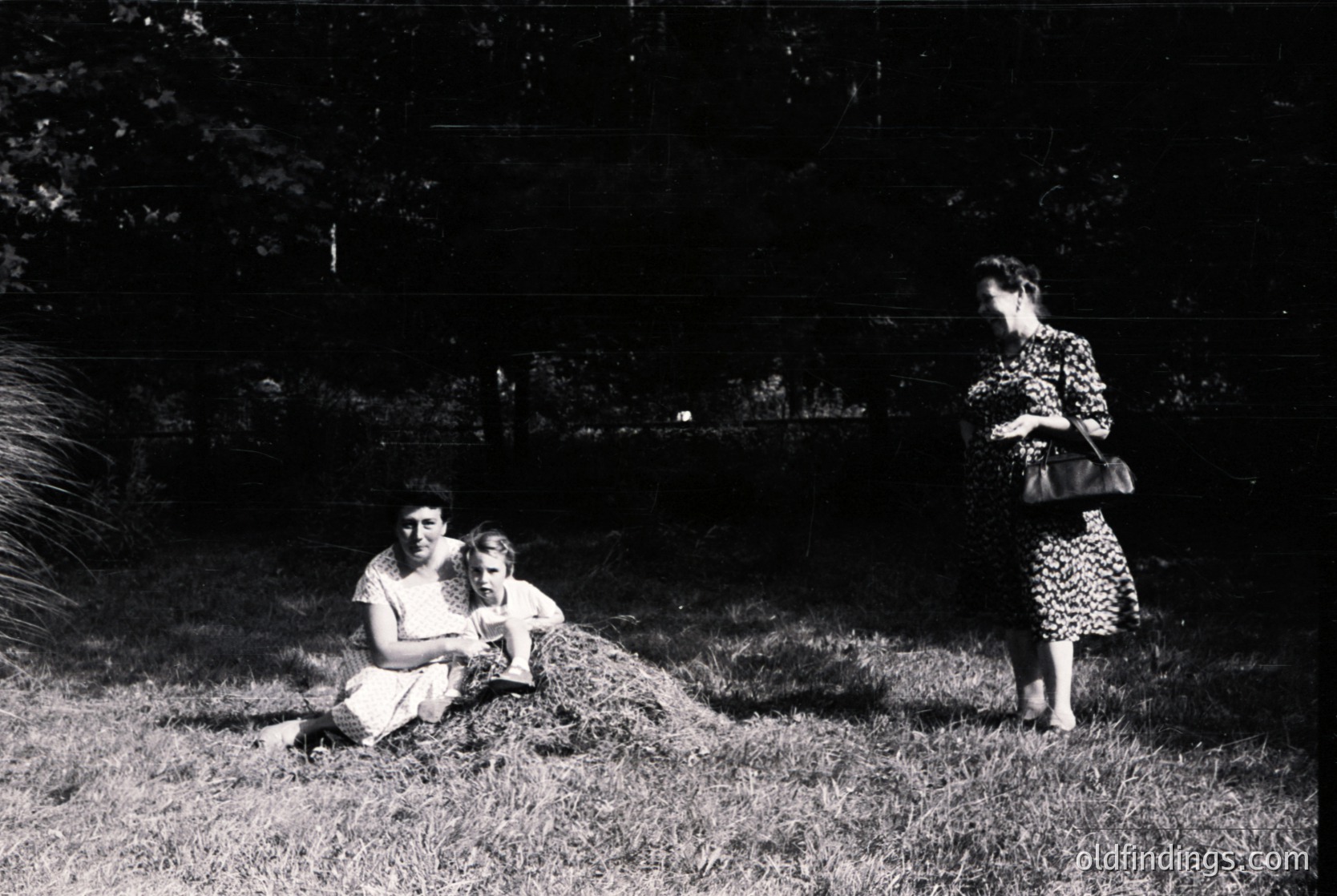 Mid-20th century black-and-white photo: two seated women (mother and child) in a grassy garden, one holding a small animal; a third woman in patterned dress stands nearby. Lush foliage and a wooden fence frame the scene, suggesting a private residential setting.