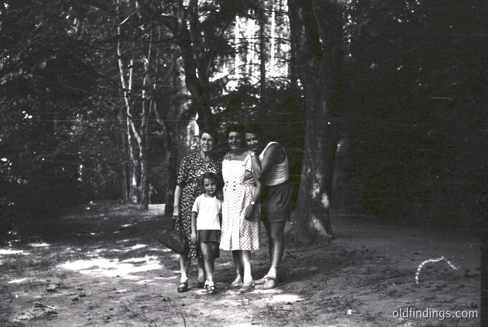 Three women and a child pose outdoors in a wooded area, likely mid-20th century. The woman on the left wears a patterned dress with a headscarf, the child a short-sleeve dress, and the woman on the right a sleeveless dress with a belt. Trees frame the scene, suggesting a park or garden setting.