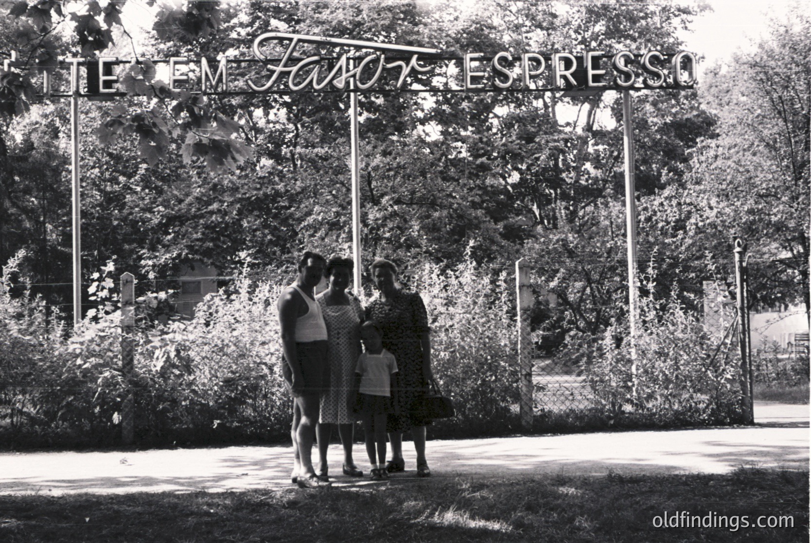 Mid-20th century black-and-white photo: Four people pose under a sign reading *"TEM FAVOR, ESPRESSO"* (Portuguese for "Please, Espresso") in a lush, tree-lined area. Sign’s cursive script and vintage typography suggest 1950s–1960s. Likely a café or espresso stand in a park or urban green space. é