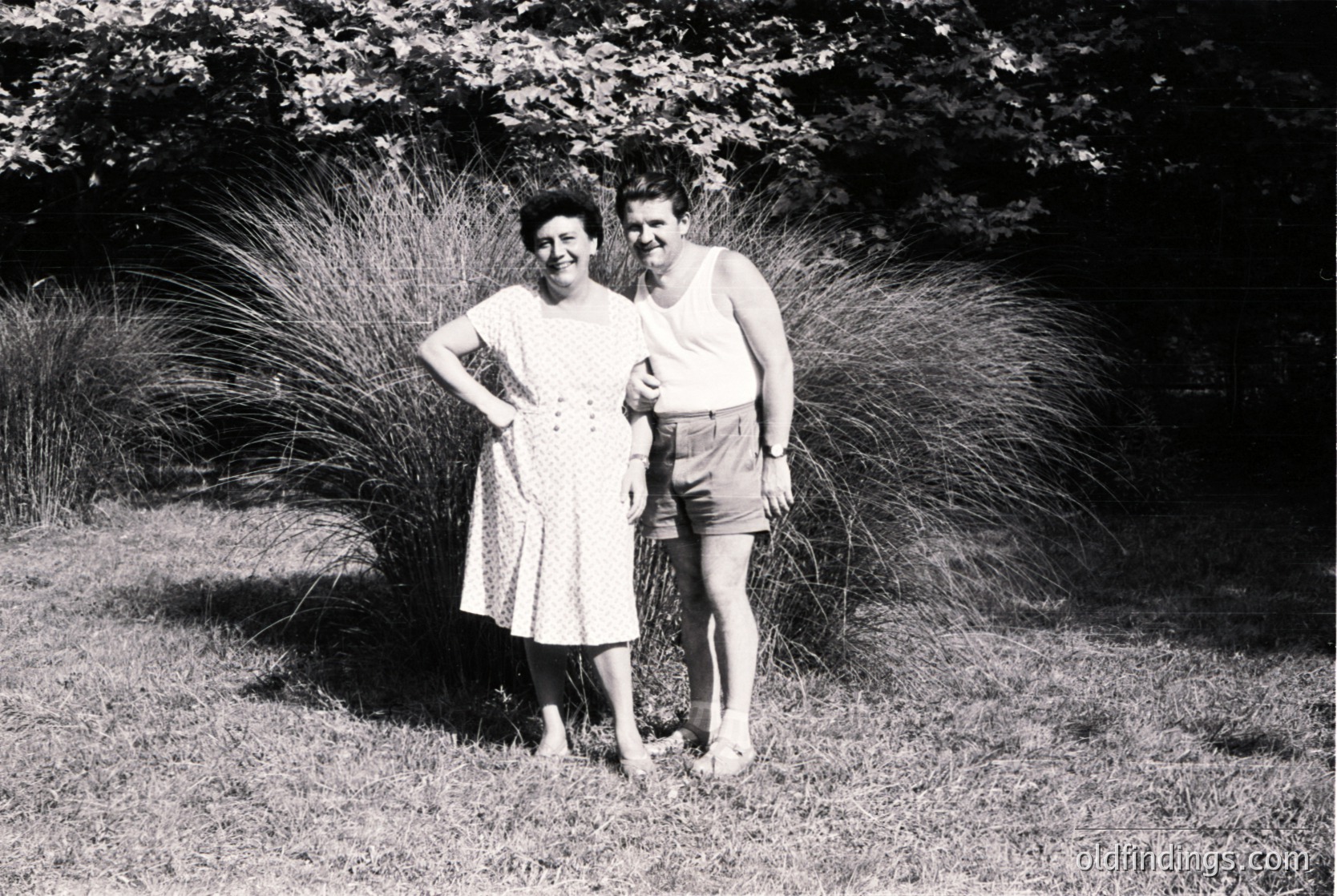 Couple posing outdoors in mid-20th century attire—woman in a knee-length floral dress, man in sleeveless shirt and shorts. Lush greenery and ornamental grasses frame them. Likely 1950s–1960s, Western Europe/USA.