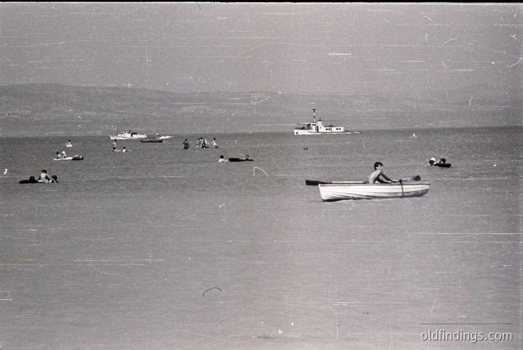 Mid-20th century seaside scene: crowded beach with swimmers, a lone rowboat, and distant boats. Worn grainy texture suggests vintage or coastal tourism. Potential or Eastern European seaside resort vibe.