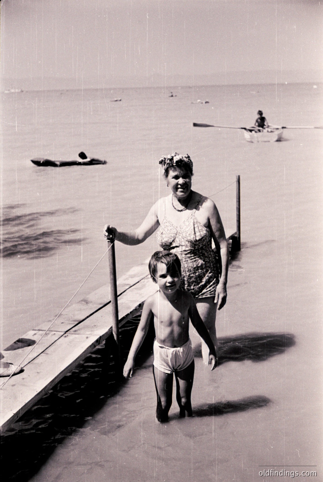 A woman and child in vintage swimwear walk along a wooden pier into shallow water, likely a seaside resort. The woman holds a long pole, and a rowboat with two figures is visible in the background. Mid-20th century beach culture, possibly Eastern European.