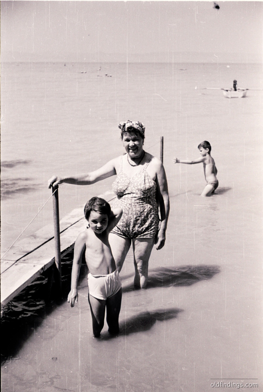 A woman in a floral swimsuit and headband leads a young boy in swim diapers along a shallow beach edge, 1950s-60s. Another child plays in the water, while a distant swimmer rests. Classic seaside family moment.