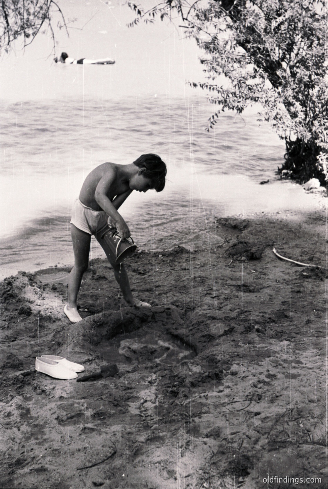 Mid-20th-century beachgoer in shorts and sandals kneels to place a small wooden box on sandy shore, near shallow water. Classic vintage seaside attire and minimalist beach setting. Likely