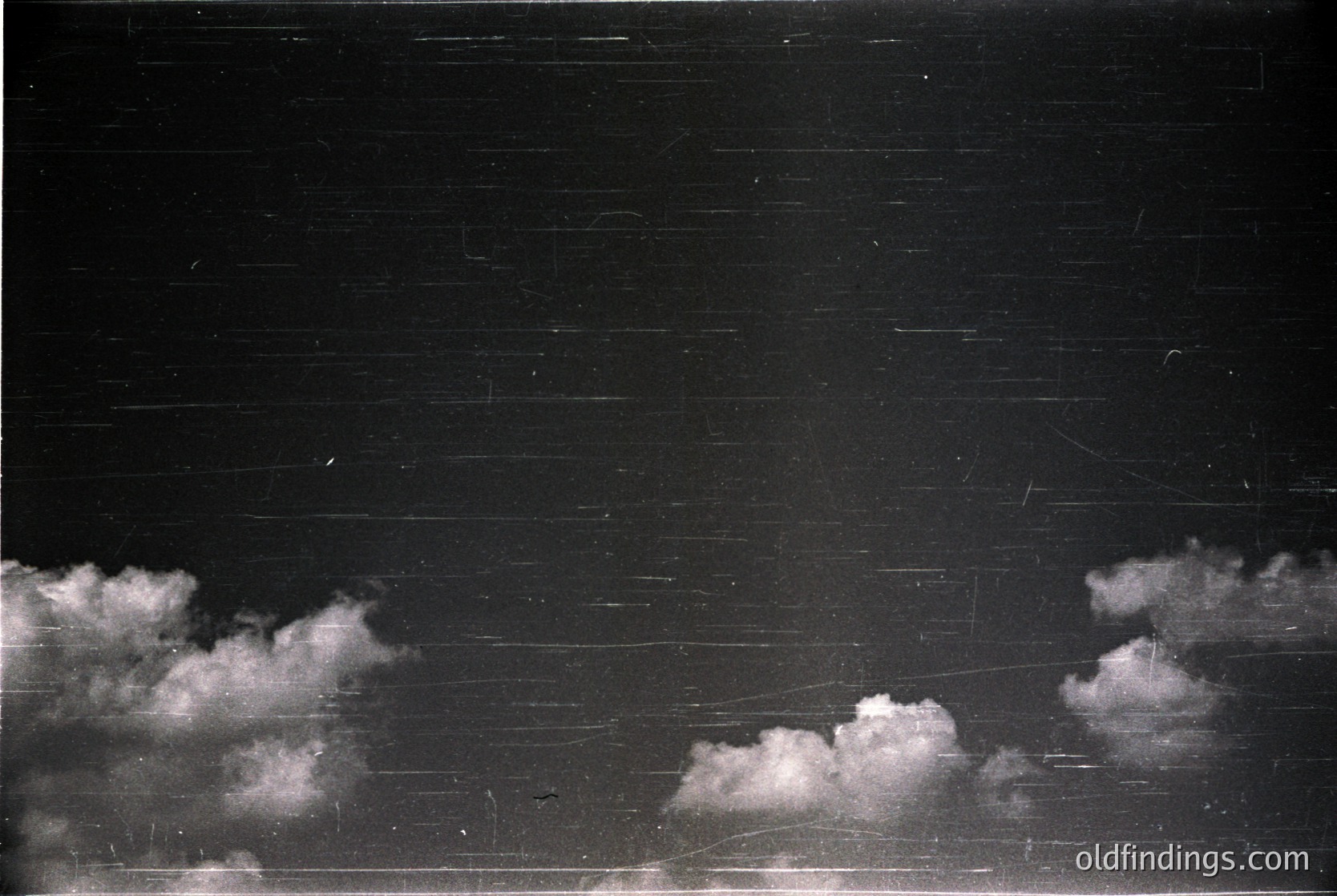 High-contrast black-and-white shot of a brick wall under dramatic cloud formations. Horizontal brickwork suggests urban architecture, likely residential or institutional. Scratches and minor imperfections on the wall indicate age and exposure. Composition emphasizes texture and light contrast.