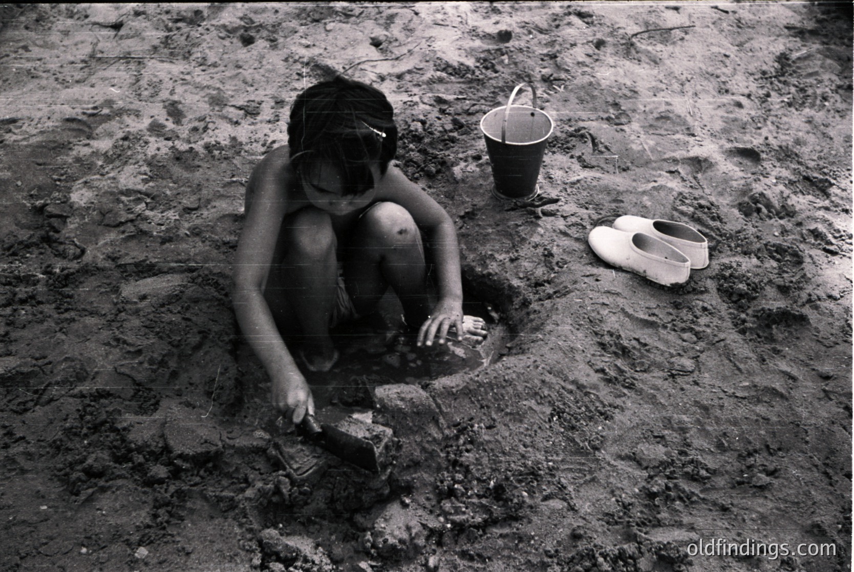Child kneeling in muddy excavation, holding small shovel. Nearby: bucket and worn sandal. Mid-20th century agricultural or construction scene, likely rural. Evokes themes of labor and childhood.