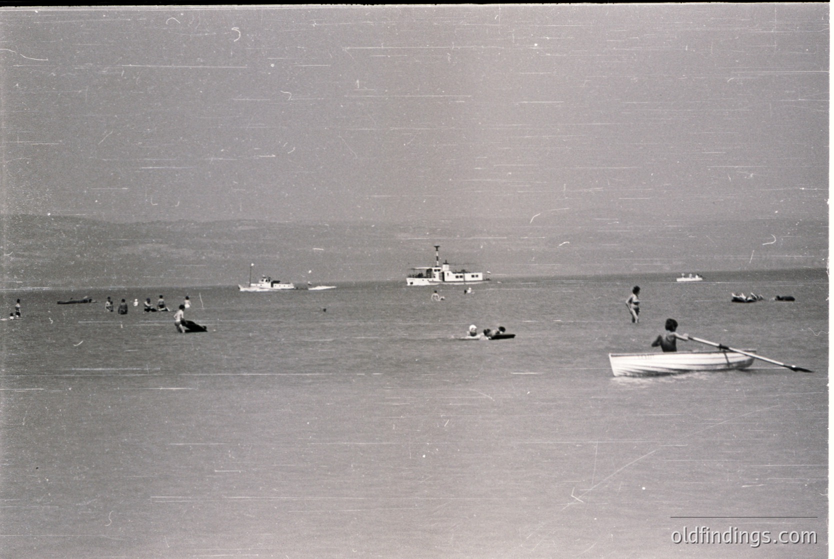 Black-and-white coastal scene featuring mid-20th century beachgoers in shallow waters. Small wooden rowboat and scattered swimmers near a distant lighthouse vessel. Flat, sandy shore with minimal vegetation. Likely Eastern European seaside resort, 1950s–1960s.