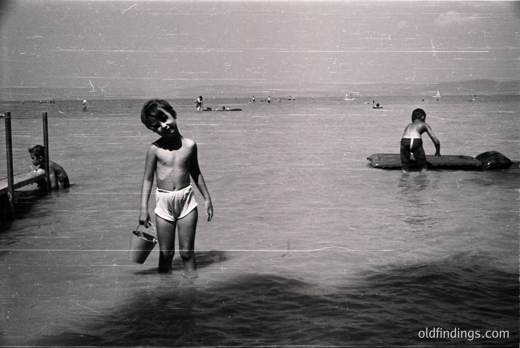 Mid-20th century seaside scene: shirtless boy in swim trunks holds a bucket near a wooden pier, wading into shallow water. Other children play in the waves and on makeshift rafts. Crowded beach with distant swimmers. Evokes midcentury coastal leisure.